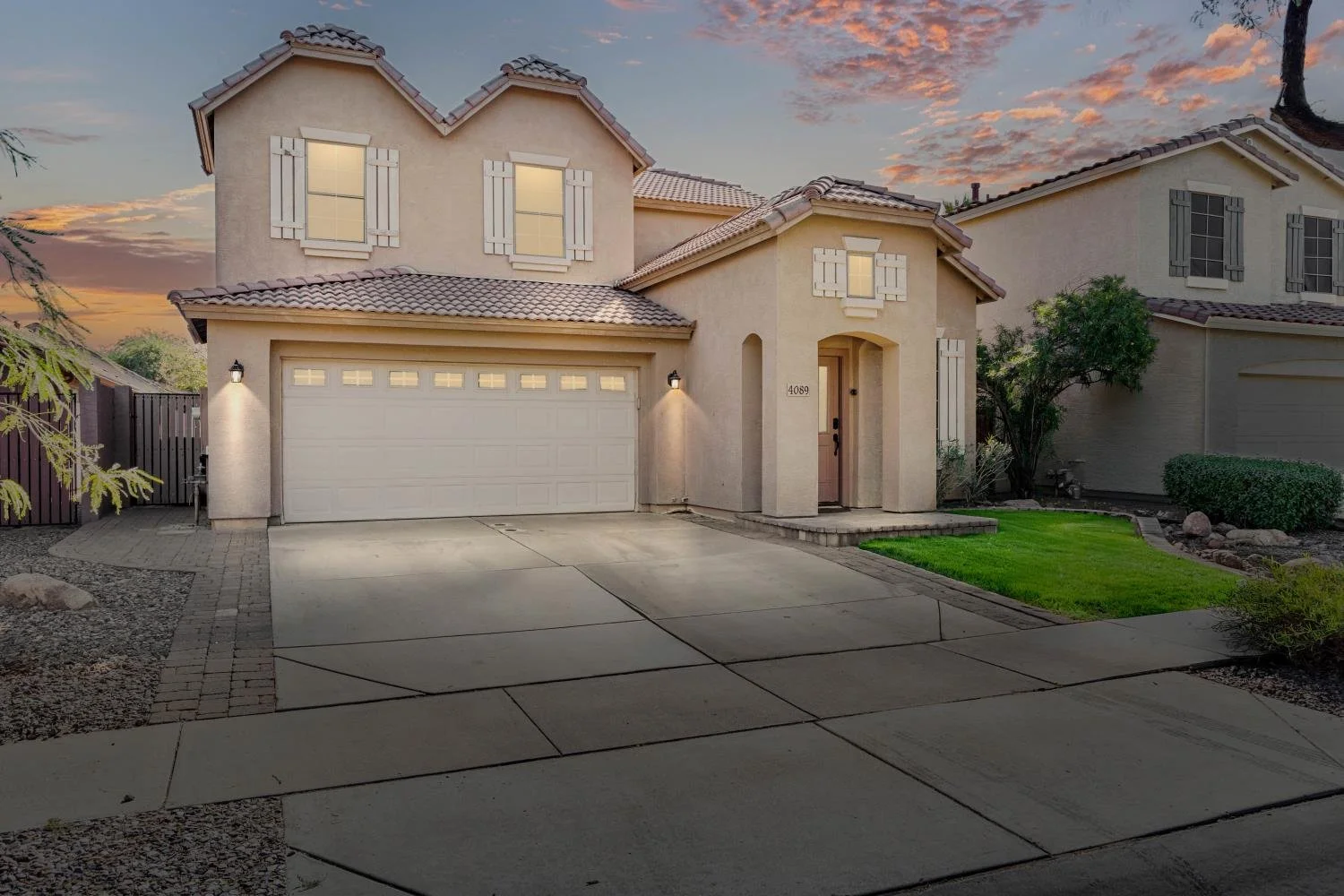 A two-story house during sunset with a beige exterior, a white garage door, a small front porch, and a well-maintained lawn with green grass and landscaping.