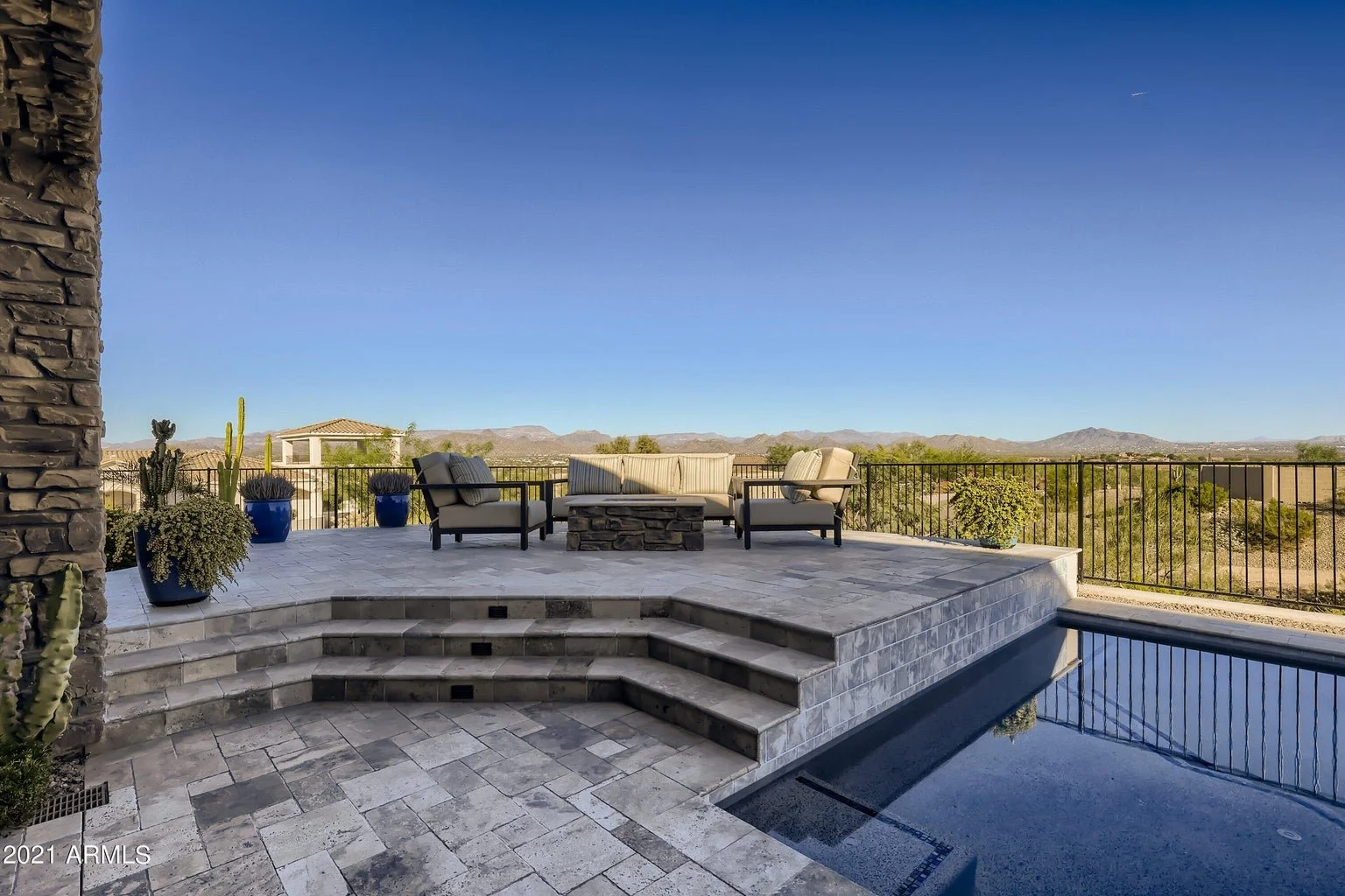 View of a luxurious backyard patio with outdoor furniture, potted cacti, and a swimming pool, overlooking a desert landscape with mountains in the background.