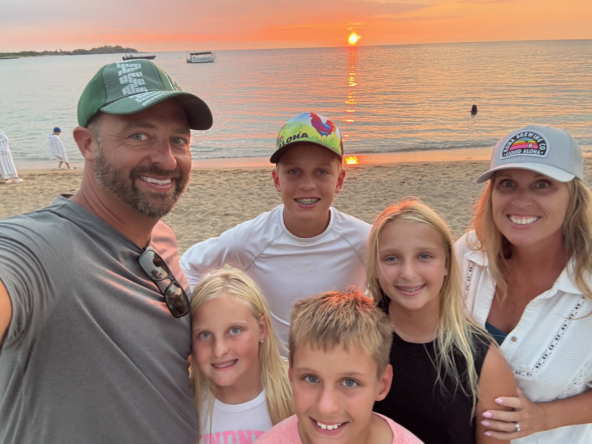 A group of six people, adults and children, smiling at the beach during sunset, with boats on the water in the background.
