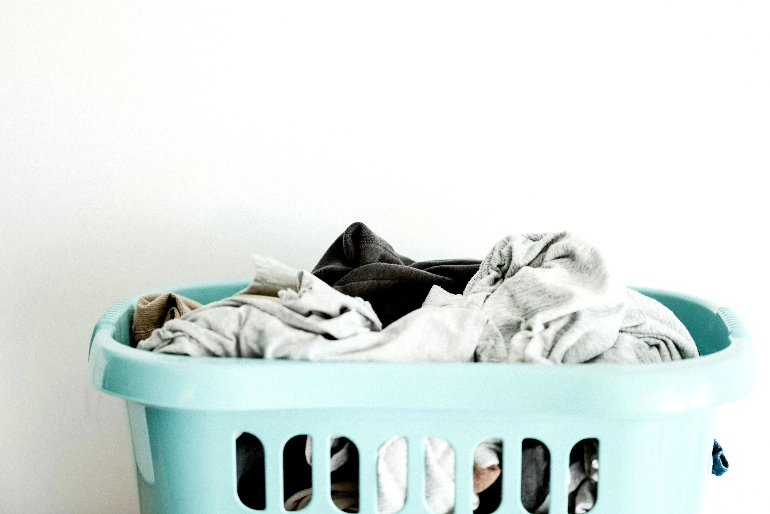 A light blue laundry basket filled with various crumpled clothes against a plain white wall.