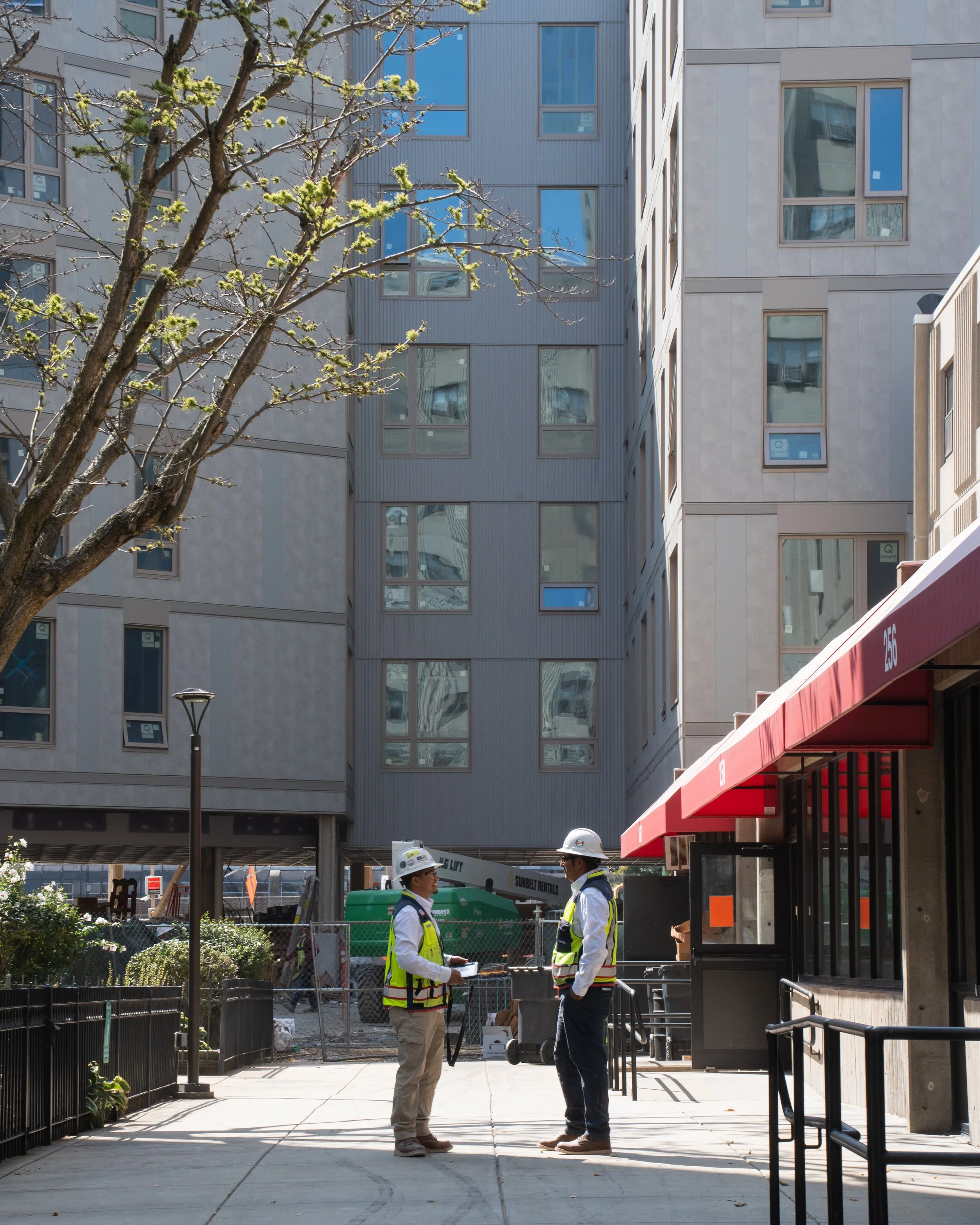 Two construction workers wearing white helmets and yellow safety vests conversing outside a building, with residential or office buildings and construction equipment in the background.