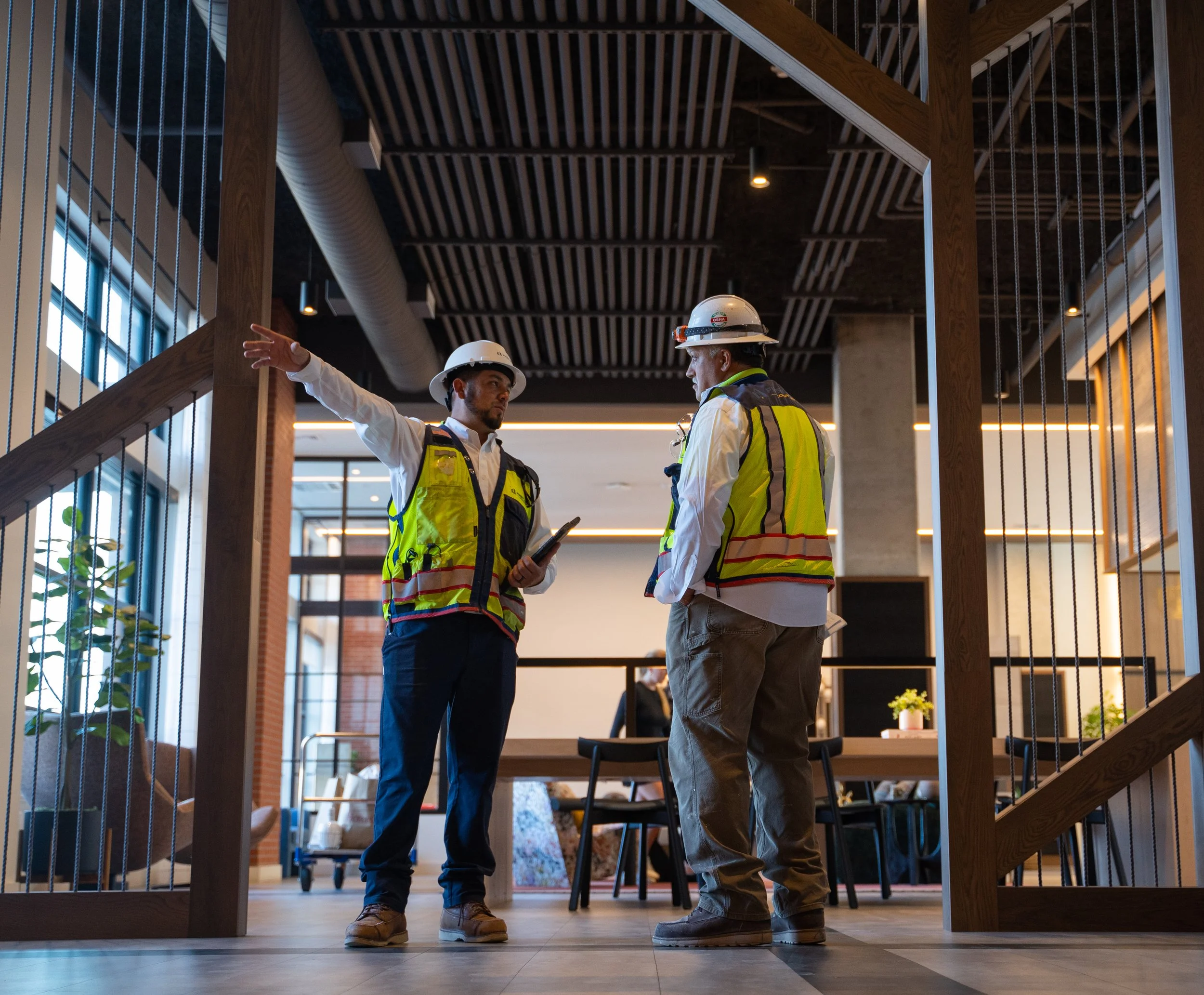 Two construction workers wearing yellow safety vests and white hard hats having a discussion indoors. One worker points to something while holding a tablet.