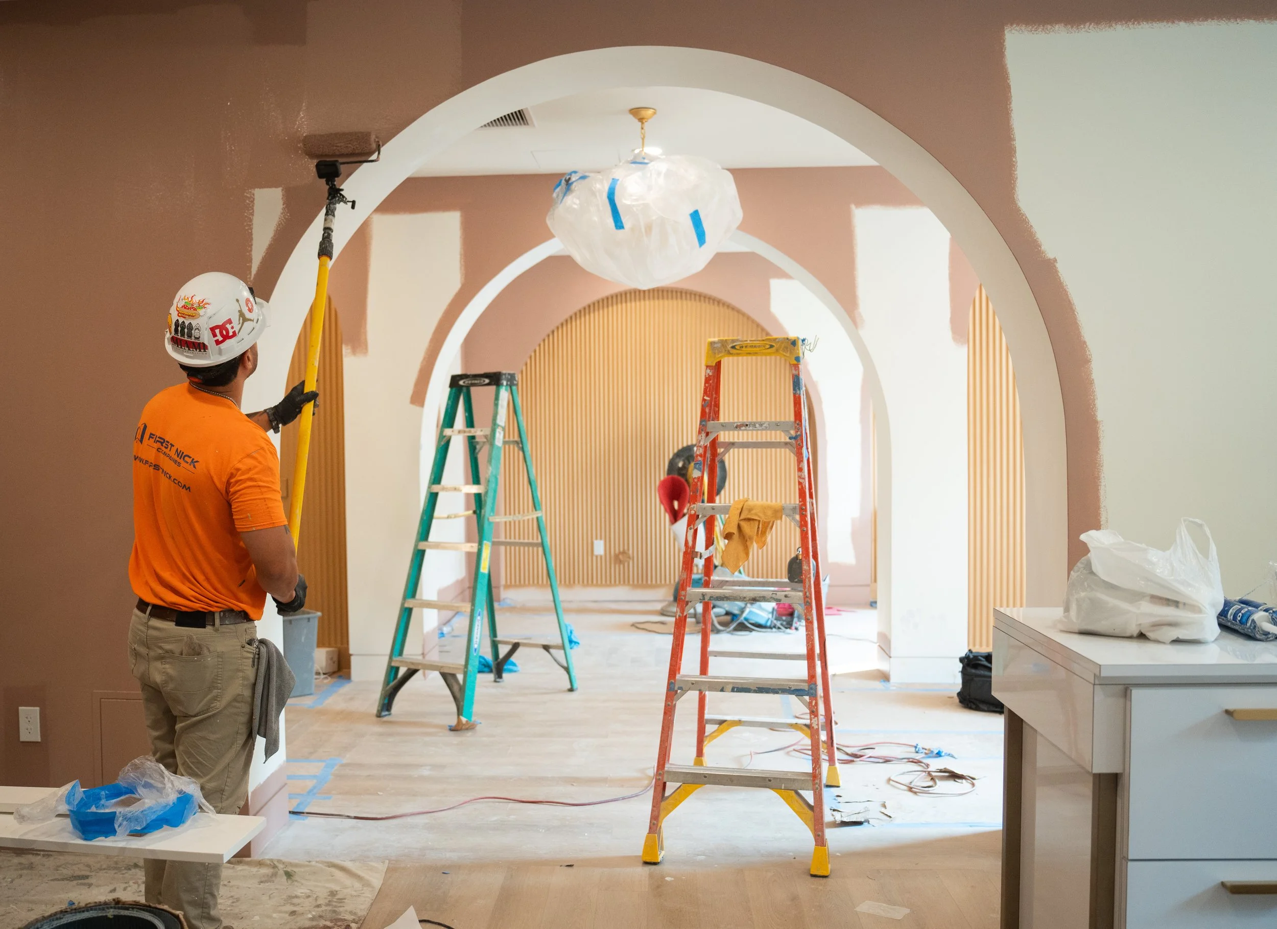 Interior of a house under renovation with workers, ladders, and painting supplies, featuring arched doorways and a chandelier covered in plastic.