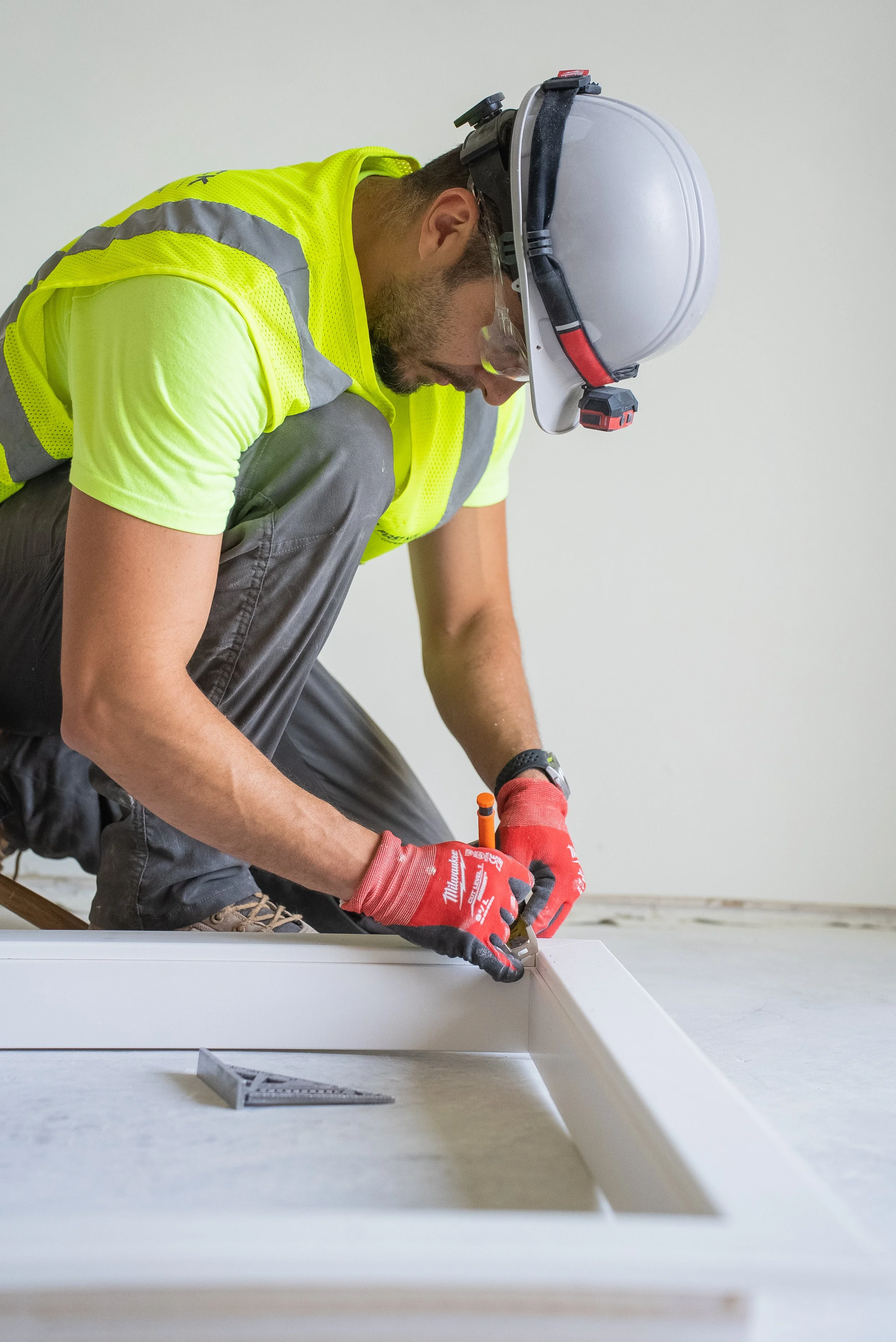 A construction worker wearing a yellow safety vest, a white helmet, and red gloves kneeling on the floor, using a tool to work on a white frame or panel.