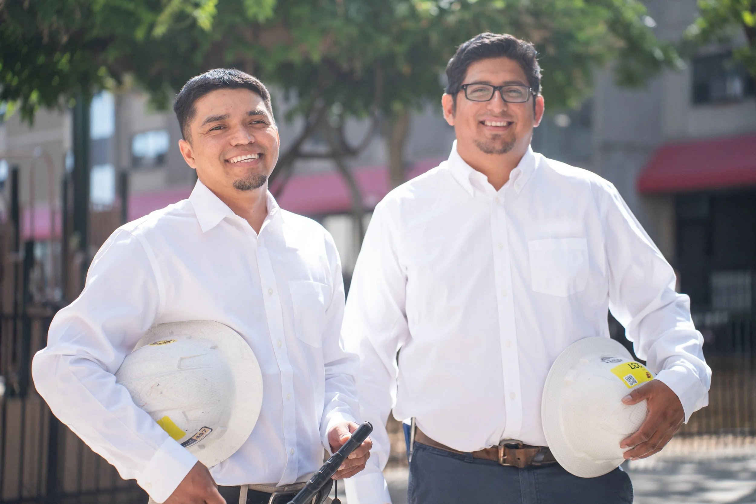 Two men in white shirts holding white helmets, standing outdoors with trees and buildings in the background.