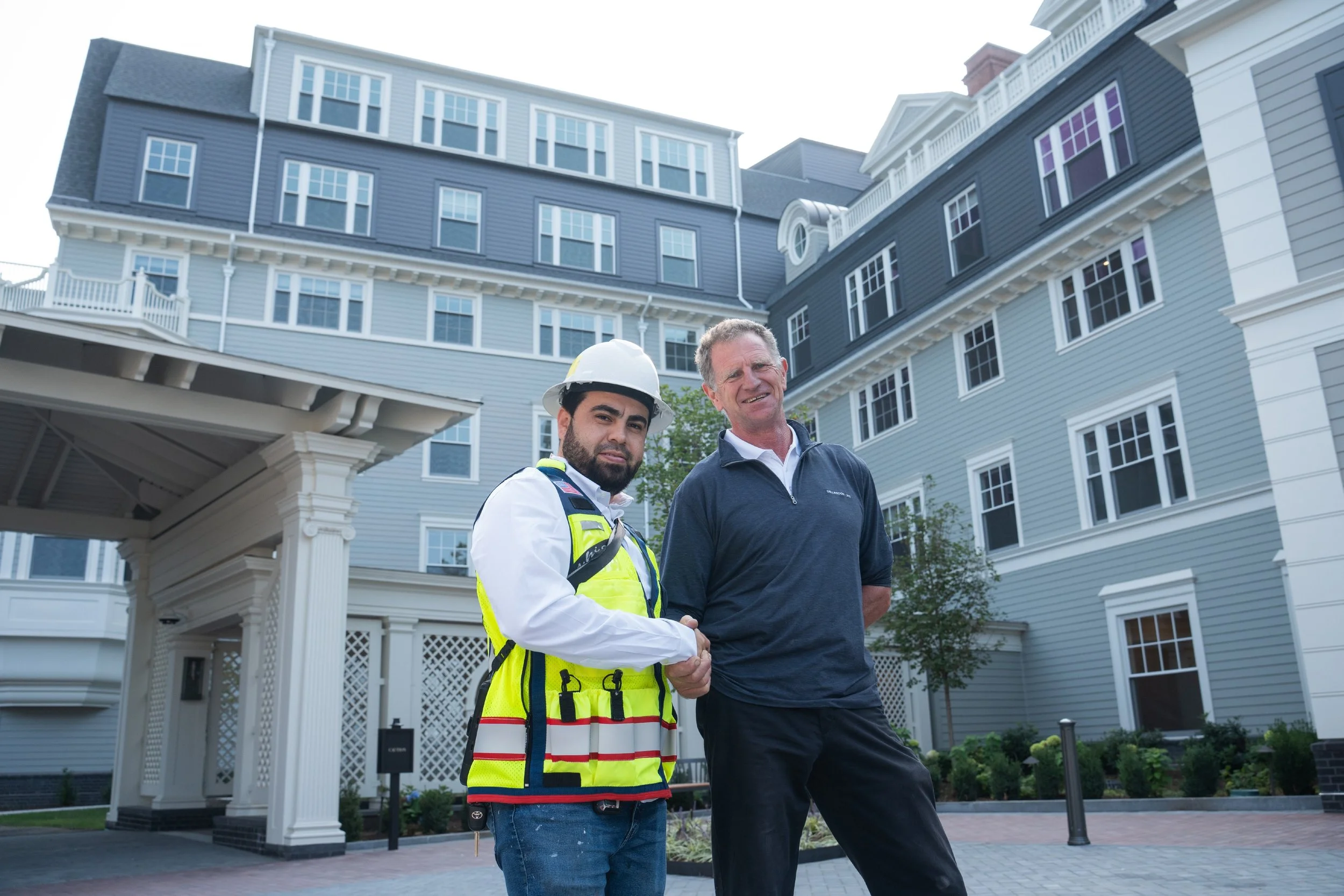 A construction worker in a high-visibility vest and helmet shaking hands with a man in a navy blue shirt in front of a large blue residential building.
