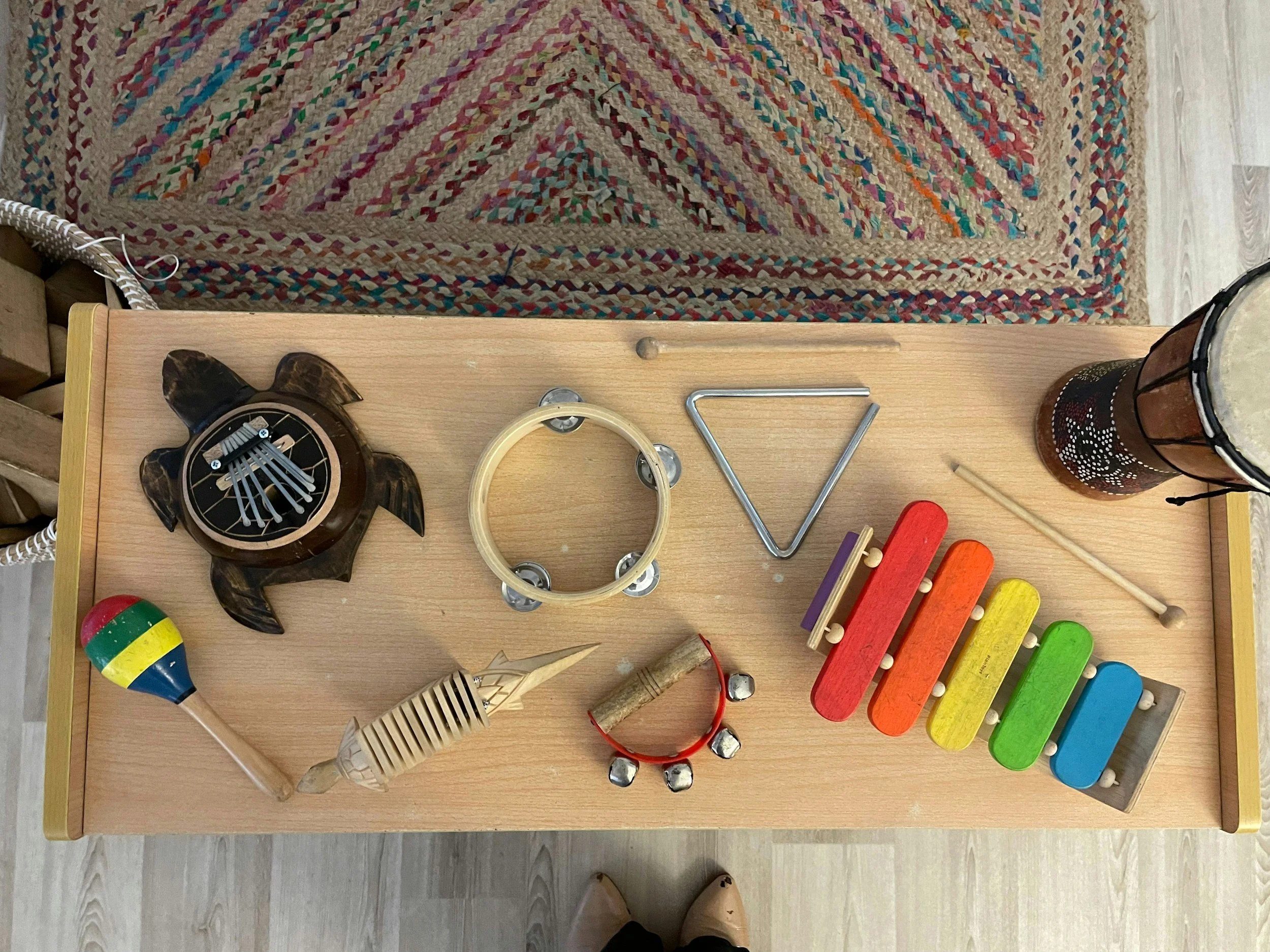 A wooden sensory table with various musical toys including a turtle-shaped percussion instrument, a tambourine, triangle, drum, rainbow-colored maraca, wooden stick, wooden xylophone, bunch of bells, and a hand drum. A woven rug with geometric patterns is in the background.