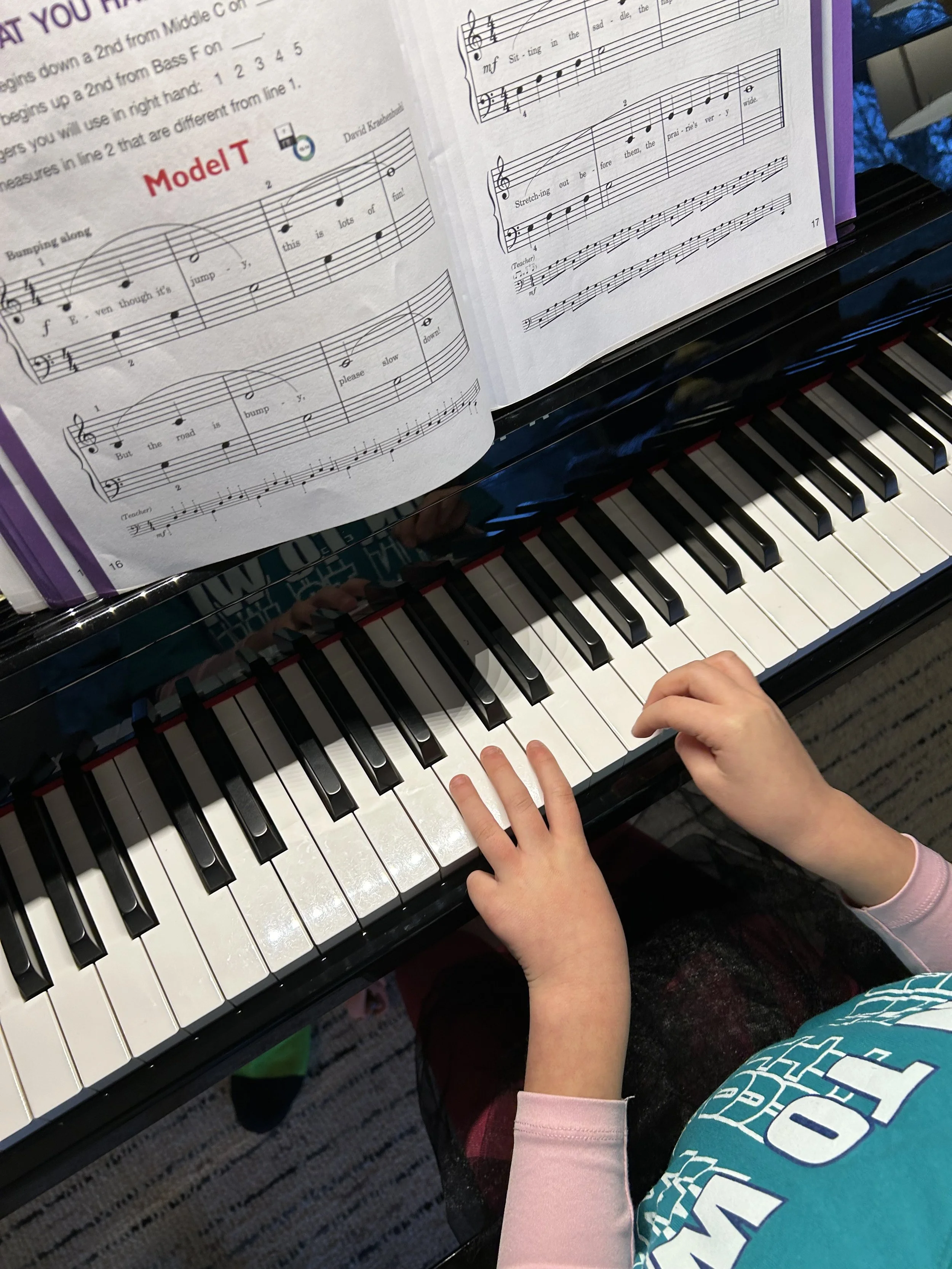 Close-up of a child's hands playing piano with sheet music open in front of them. The sheet music shows musical notes and lyrics. The child is wearing a teal shirt with white text and a pink long-sleeve shirt underneath.