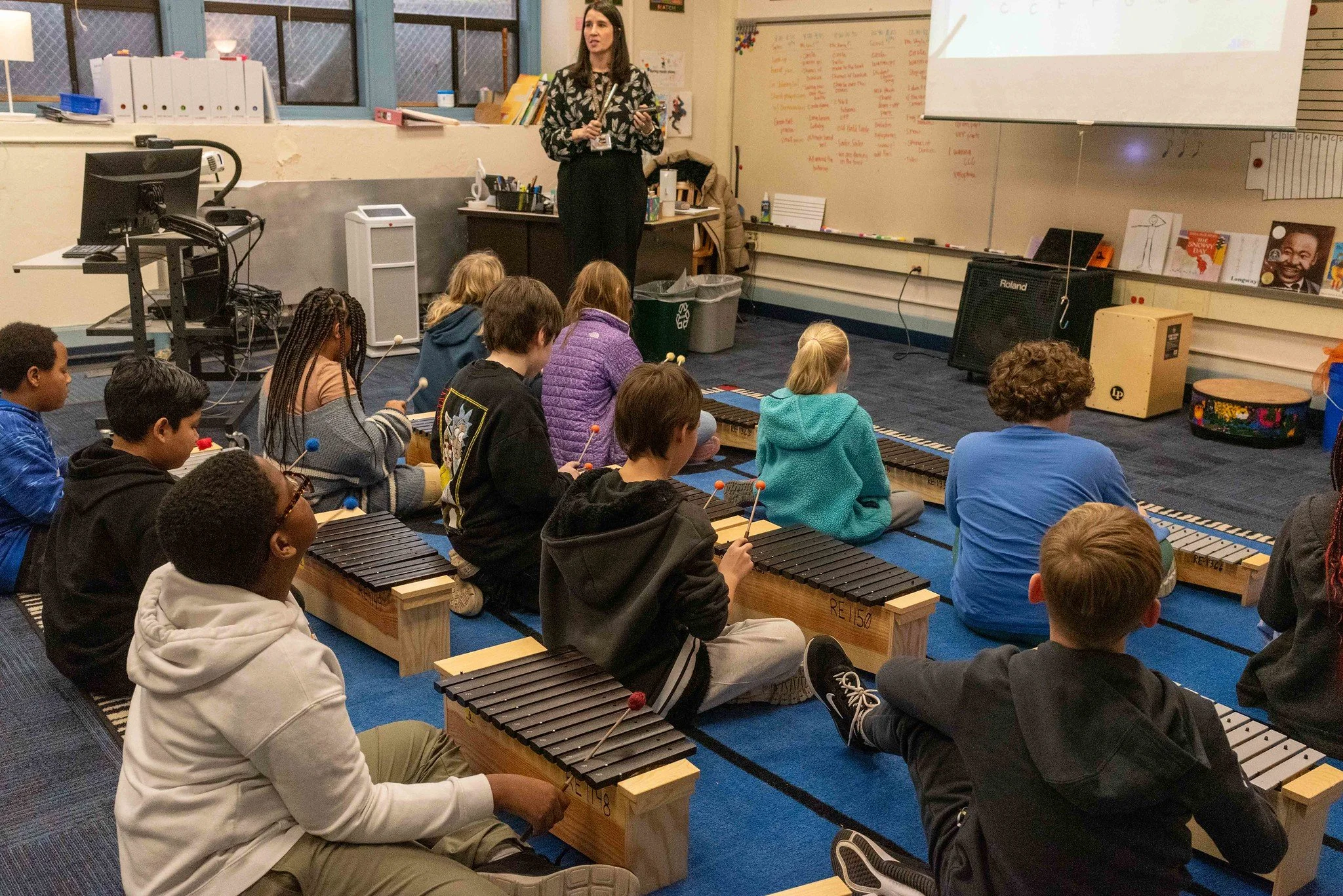 Classroom with students sitting on the floor playing xylophones, facing a teacher at the front of the room.