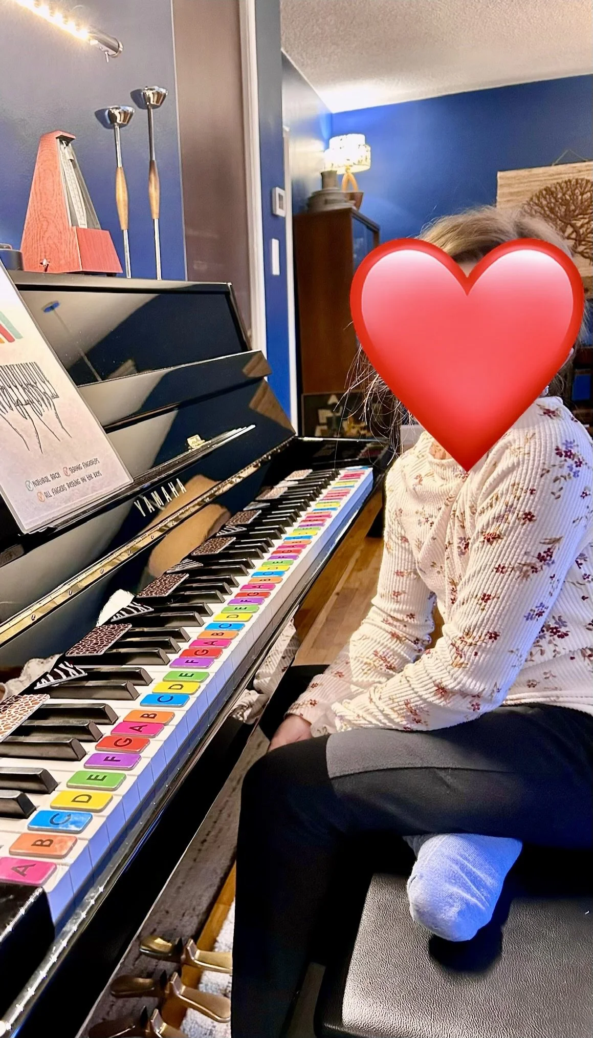 A child with a large red heart covering their face, wearing a floral shirt and sitting at a Yamaha piano with colorful alphabet stickers on the keys.