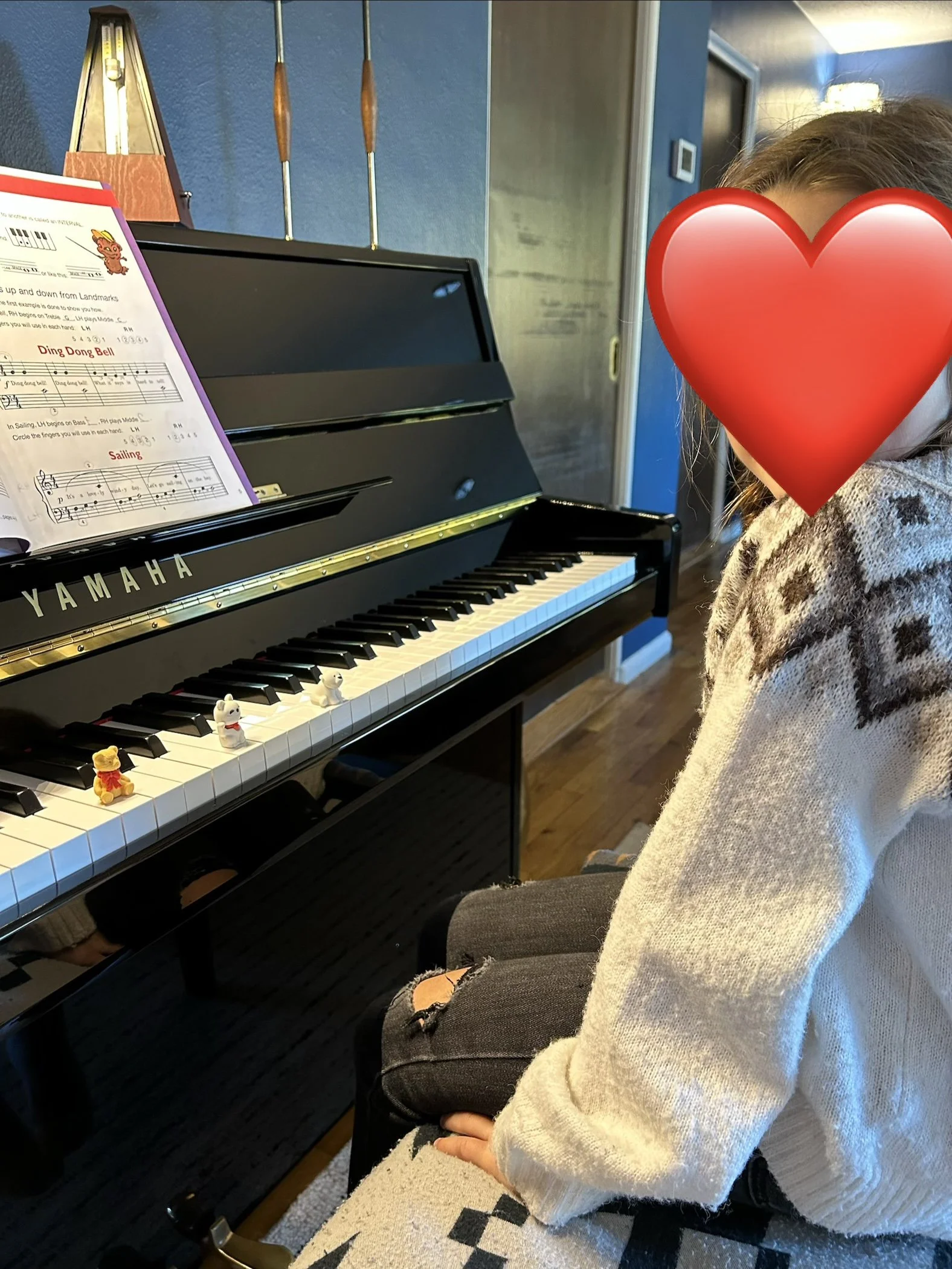 A person sitting at a black Yamaha piano with sheet music on the stand. Their face is covered with a large red heart emoji. The person is wearing a beige knitted sweater and ripped black jeans. The piano has small figurines of animals placed on the keys. In the background, there is a blue wall with some decorative items.