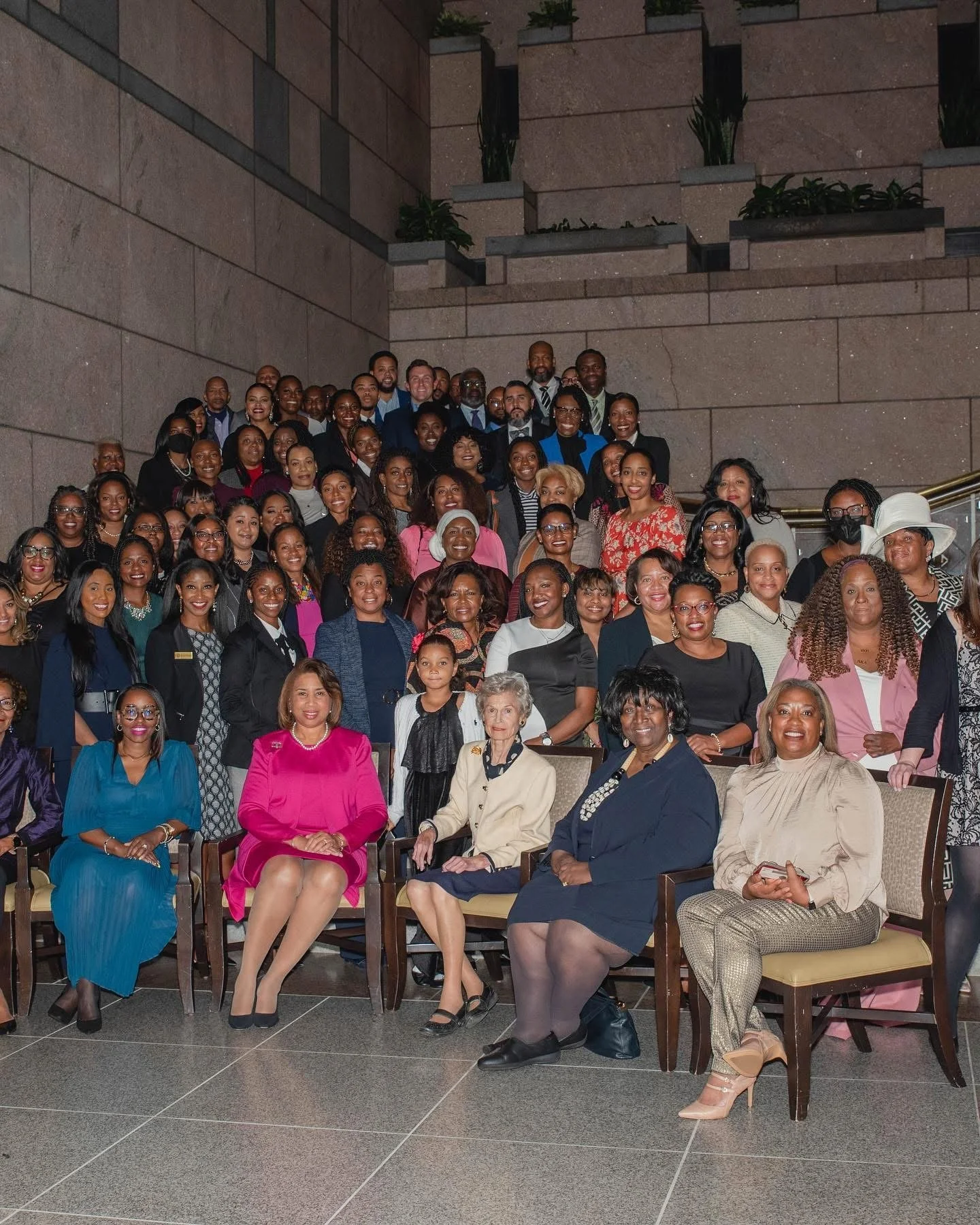 A large group of diverse women and men gathered in a spacious indoor venue, posing for a group photo on stairs and seated in front of a stone wall.