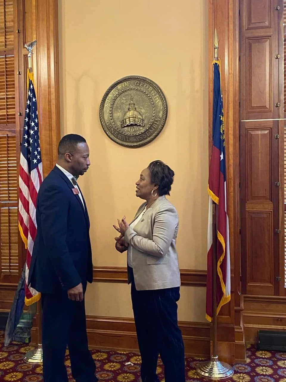 Two people, a man and a woman, standing face to face and engaging in conversation inside a wood-paneled room with flags and the Georgia state seal on the wall.