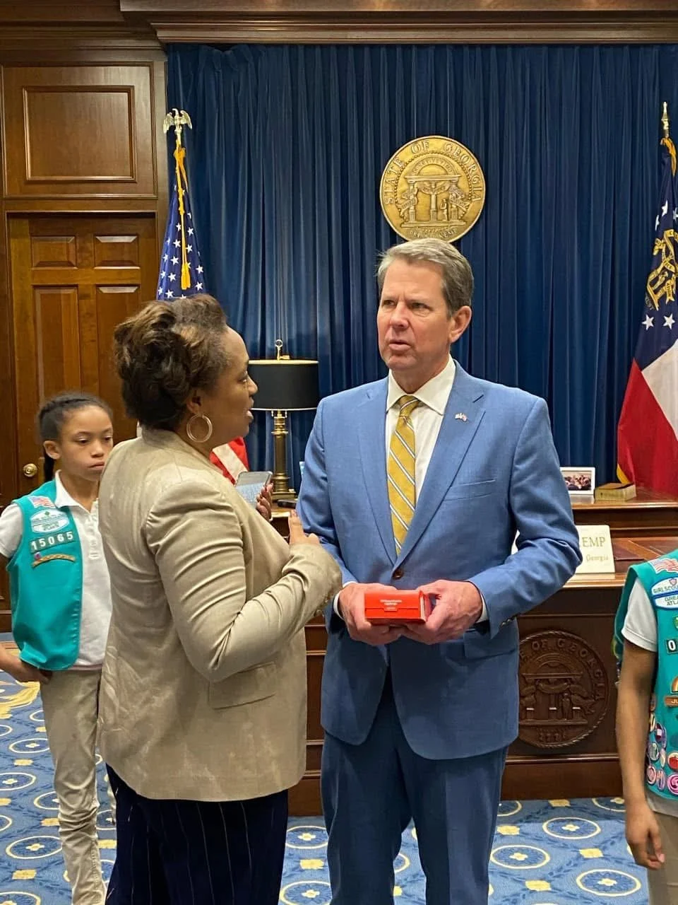 A woman and a man having a serious conversation in a formal office with the Georgia state seal on the wall behind them. The woman is wearing a beige blazer and the man is dressed in a blue suit, holding a red box. Two children in Cub Scouts uniforms 