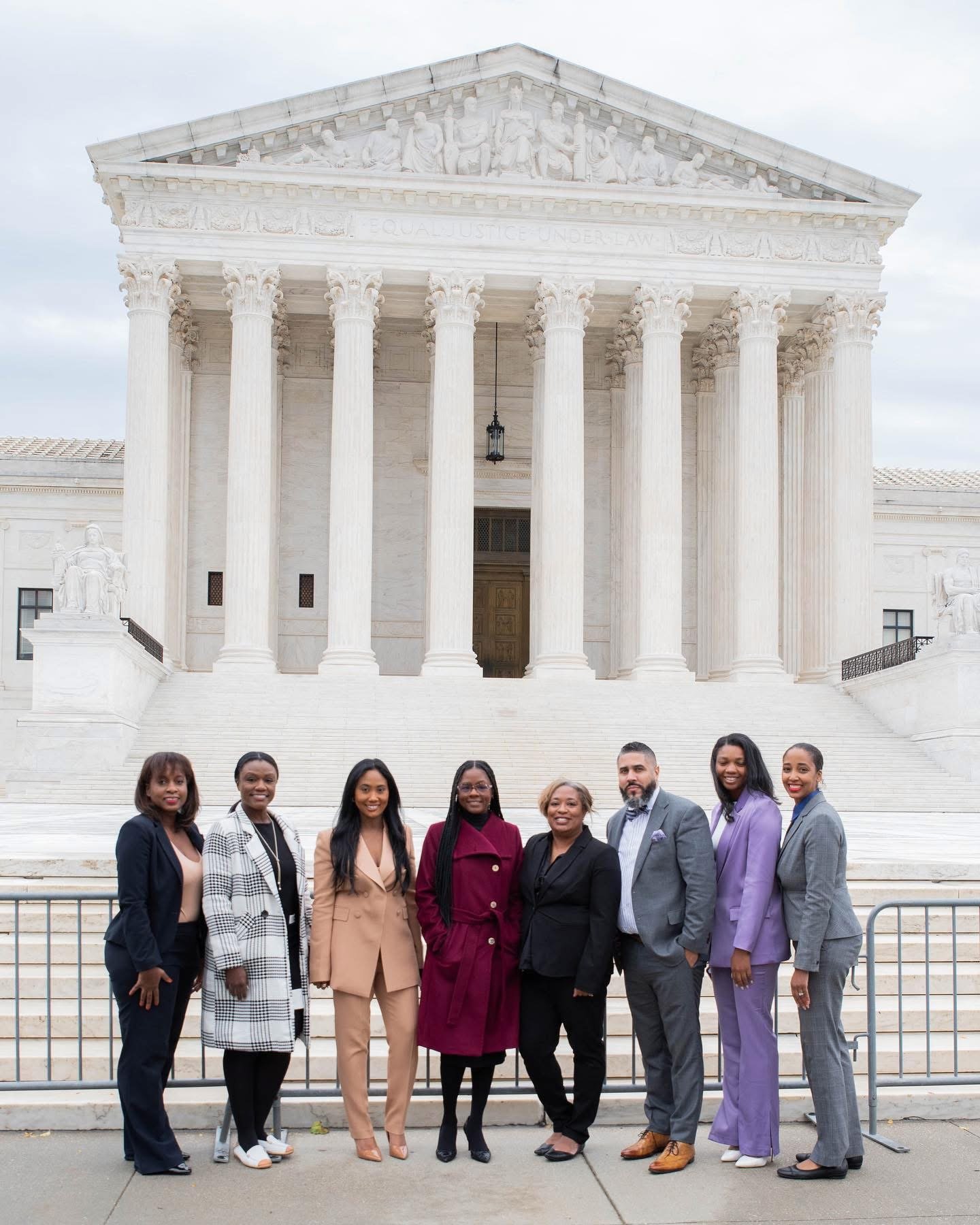 Group of eight diverse professionals standing in front of the U.S. Supreme Court building in Washington, D.C., dressed in business attire, smiling for the photo.