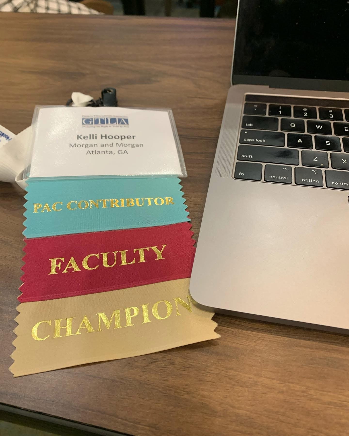 A name badge, colorful ribbons labeled 'PAC CONTRIBUTOR,' 'FACULTY,' and 'CHAMPION,' a tissue, and a partially visible laptop on a wooden table.