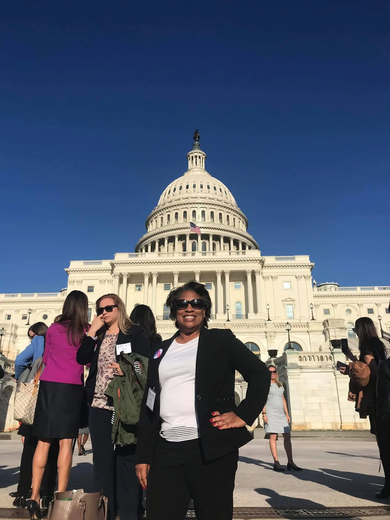 A woman smiling and wearing sunglasses in front of the U.S. Capitol building with other people in the background.