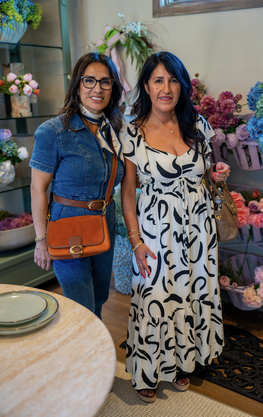 Two women standing inside a flower shop, smiling at the camera, with shelves of flowers and floral arrangements in the background.