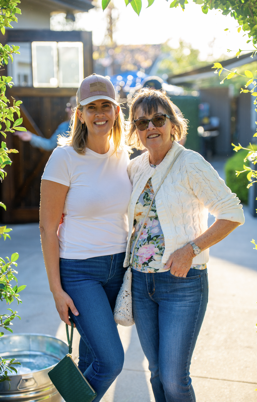 Two women smiling outdoors in a backyard, standing close together in bright sunlight, with plants and a shed in the background.
