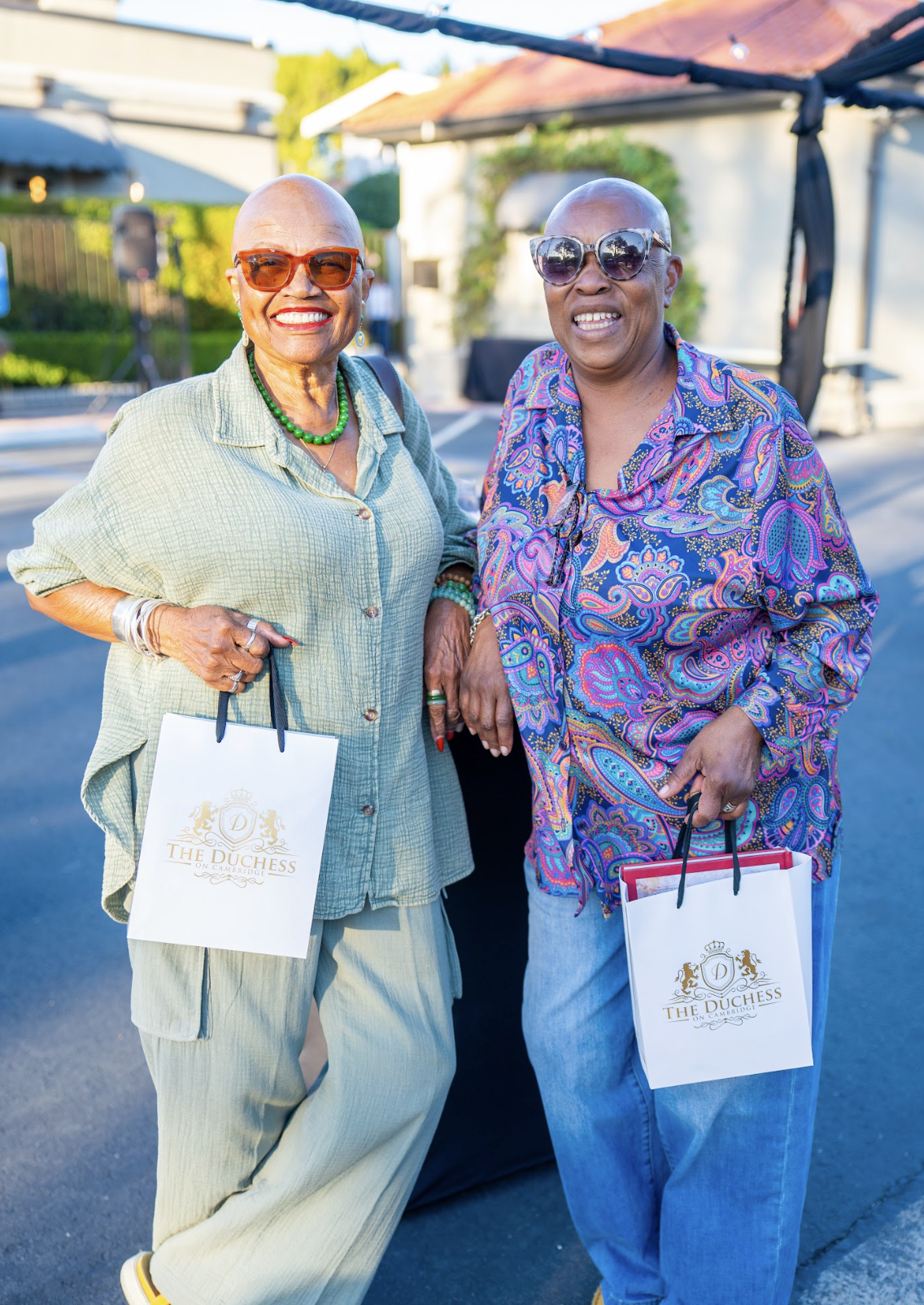 Two smiling older women wearing sunglasses, holding shopping bags, standing outdoors on a sunny day.