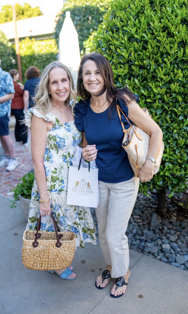 Two smiling women outdoors, standing in front of a green hedge, one holding a gift bag labeled 'The Duchess' and a straw handbag, and the other holding a beige purse and wearing a blue top and cream pants.