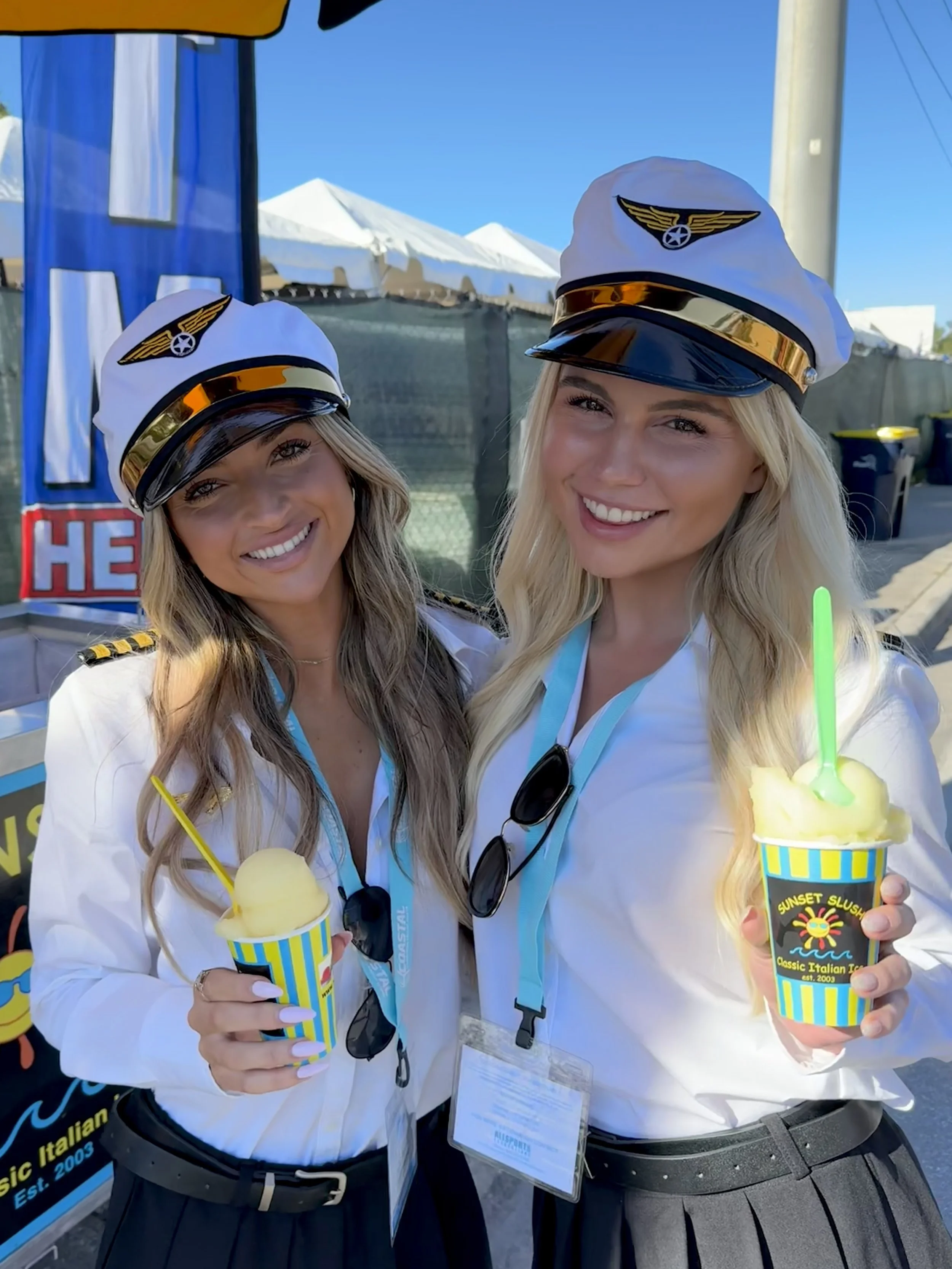 Two women in pilot costumes smiling and holding cups of gelato at an outdoor event.