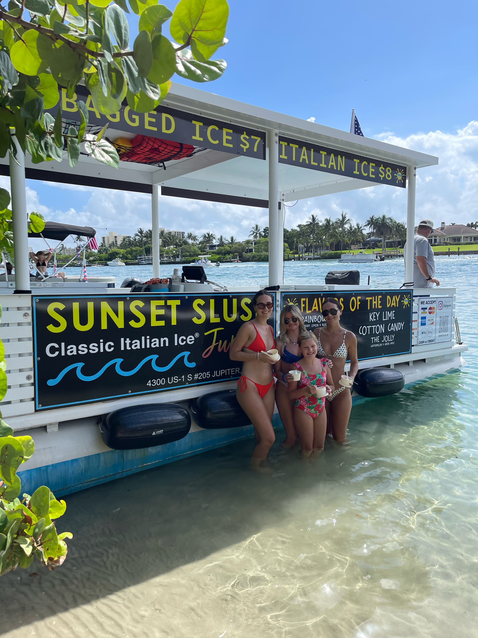 Four women in swimsuits standing in shallow water near a boat with signs advertising Italian ice, sunny sky, and palm trees in the background.
