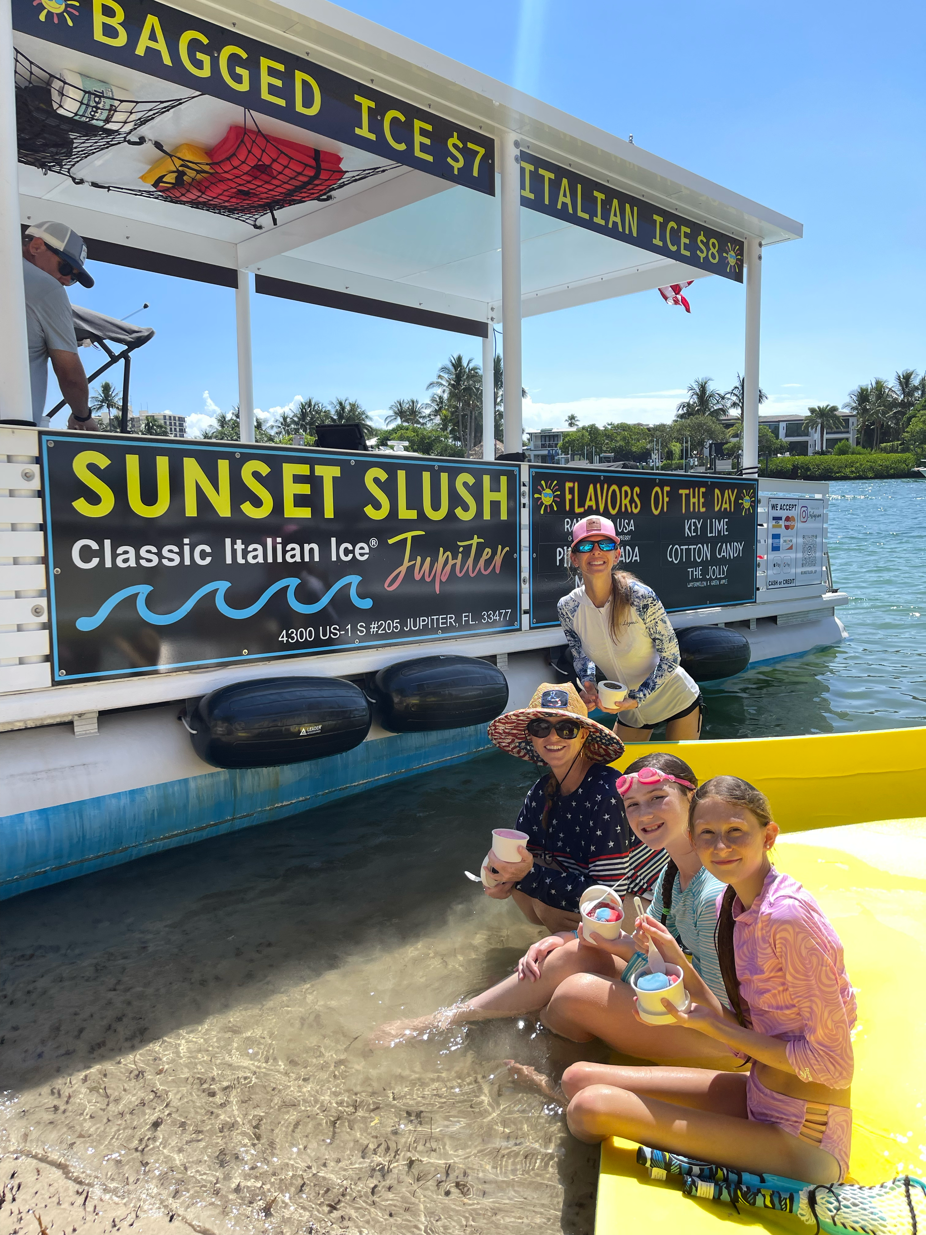 A floating ice cream stand named Sunset Slush in Jupiter, Florida, with four women and a young girl sitting on the beach and enjoying ice cream. The stand offers Italian ice flavors and has a sign displaying prices and flavors.