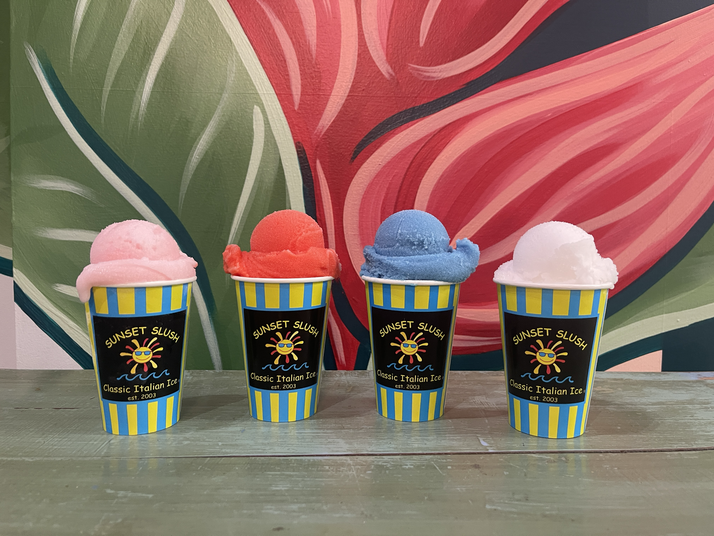 Four cups of colorful ice cream with scoops of pink, red, blue, and white, each in a yellow and blue striped cup labeled 'Sunset Slush Classic Italian Ice' on a wooden surface with a colorful abstract mural in the background.