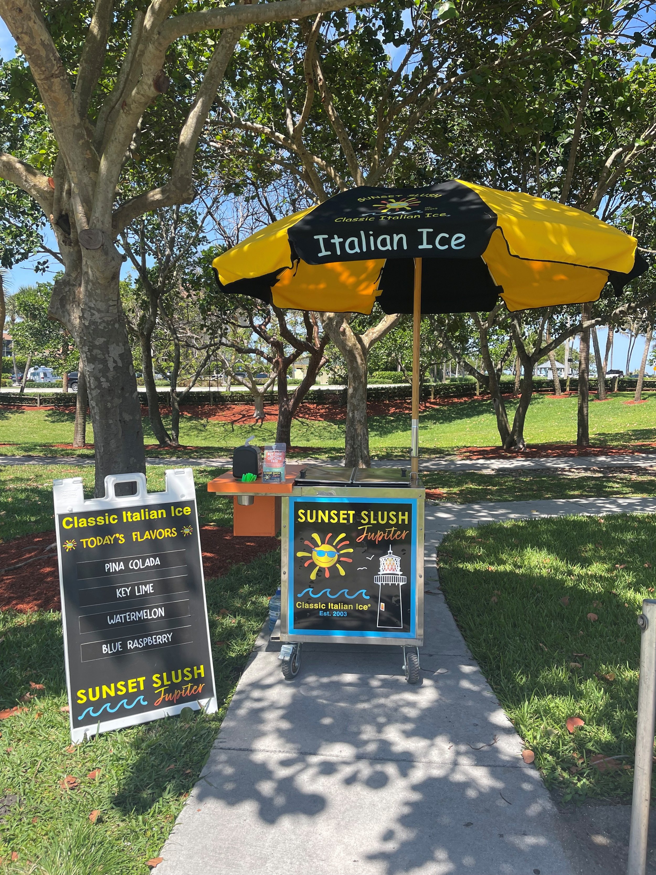 A small outdoor ice cream stand with a yellow and black umbrella that says 'Italian Ice'. There is a sign listing flavors such as Pina Colada, Key Lime, Watermelon, and Blue Raspberry. The stand has a colorful sign that reads 'Sunset Slush Jupiter' and depicts a smiling sun wearing sunglasses and a lighthouse. The stand is located on a sidewalk with grass, trees, and a park in the background.