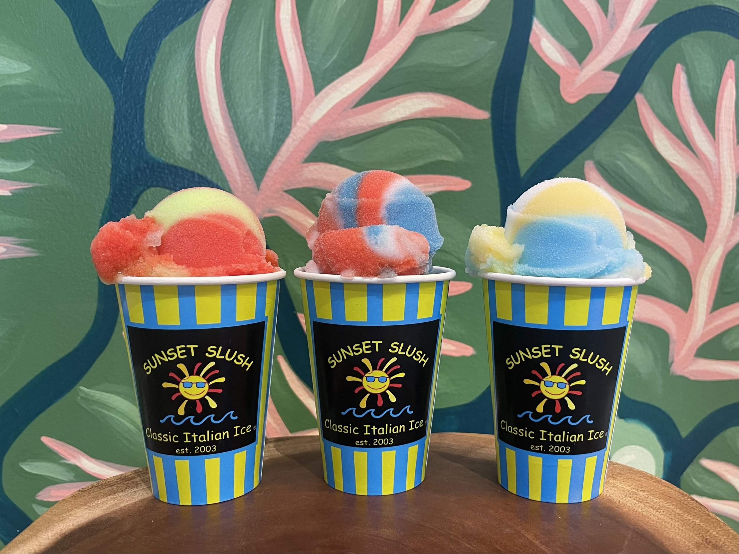 Three cups of colorful Italian ice with different flavors on a wooden surface in front of a tropical leaf patterned background.