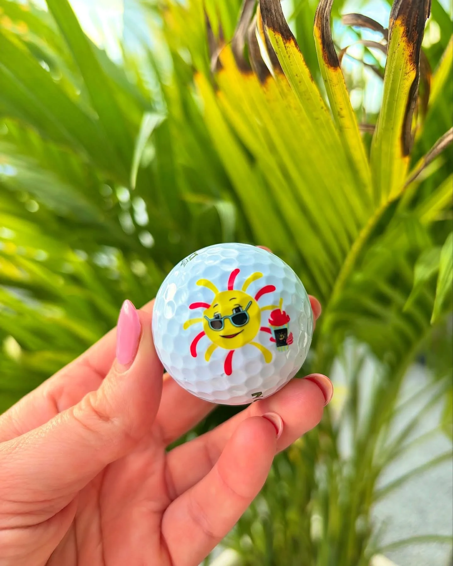Close-up of a hand holding a golf ball with a cartoon sun wearing sunglasses, holding a soft drink, with lush green plants in the background.