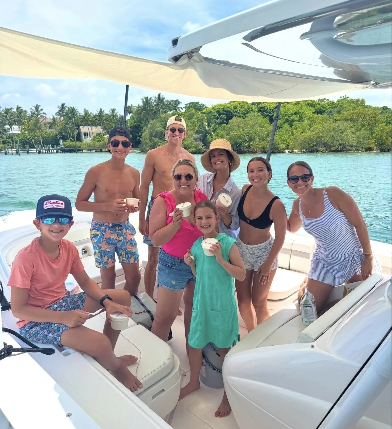 Group of people on a boat in a tropical location, smiling and holding cups, with water and lush greenery in the background.