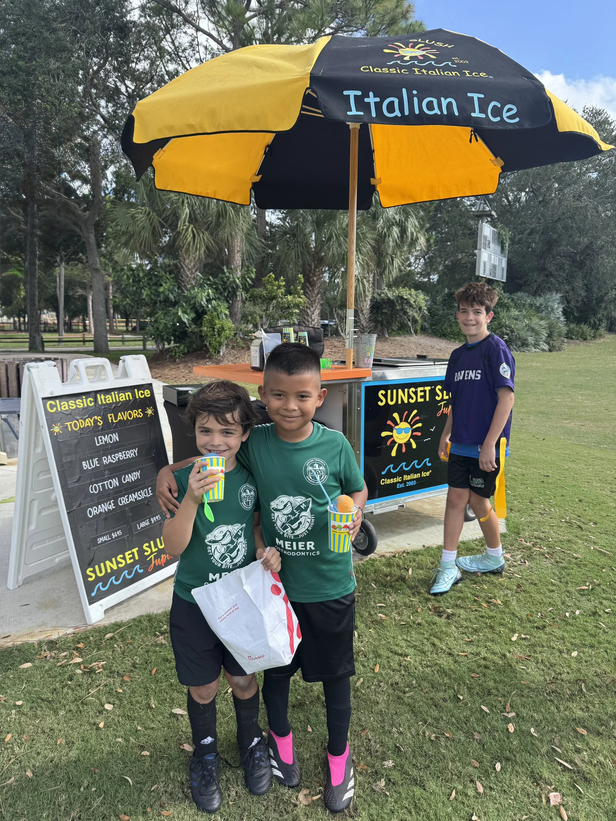 Two boys wearing green shirts standing in front of an ice cream stand, smiling and holding cups of ice cream. A third boy in a purple shirt is near the stand, smiling. The stand has a yellow and black umbrella with the words "Italian Ice" and is set outdoors with trees and grass in the background.