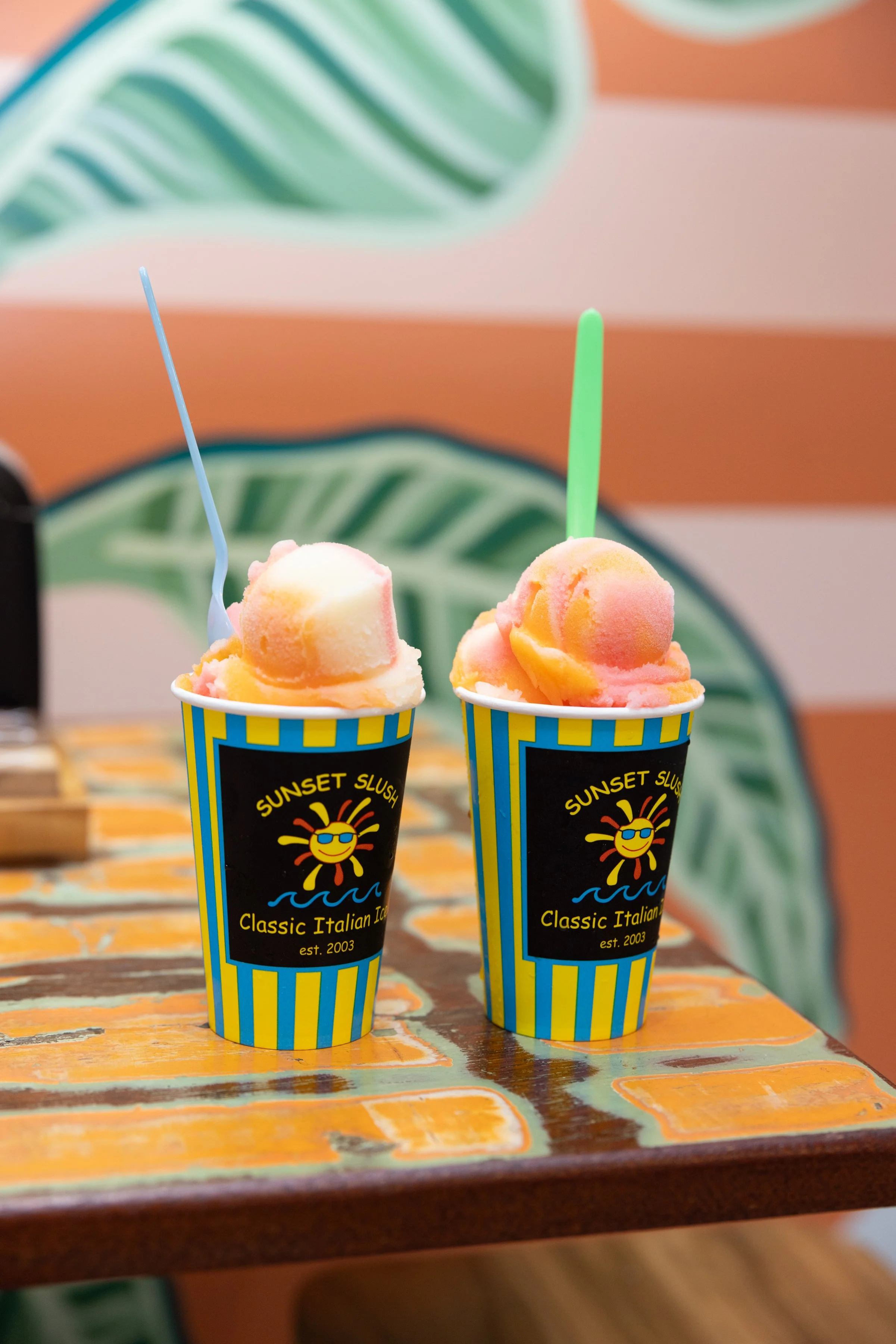 Two cups of colorful ice cream on a wooden table, with the cups labeled 'Sunset Slush' and 'Classic Italian Ice' with a smiling sun logo.