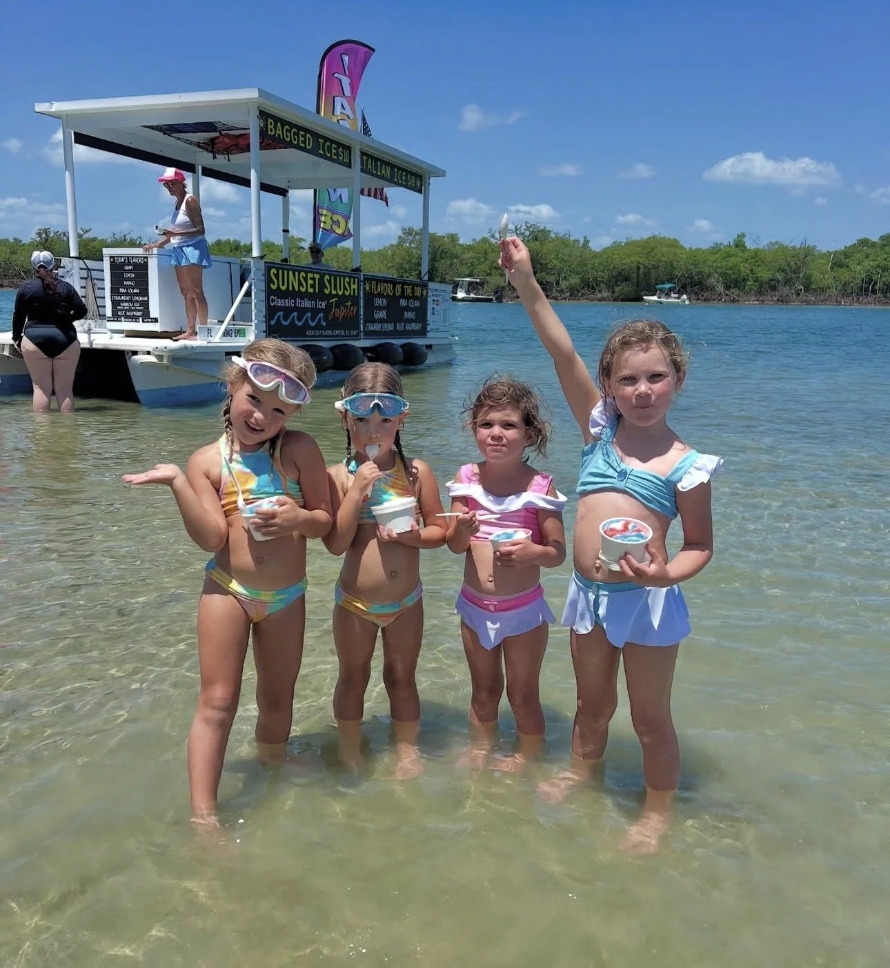 Four young girls are standing in shallow water at a beach, holding cups of ice cream and wearing swimsuits and goggles, with a boat and a floating ice cream stand in the background on a sunny day.