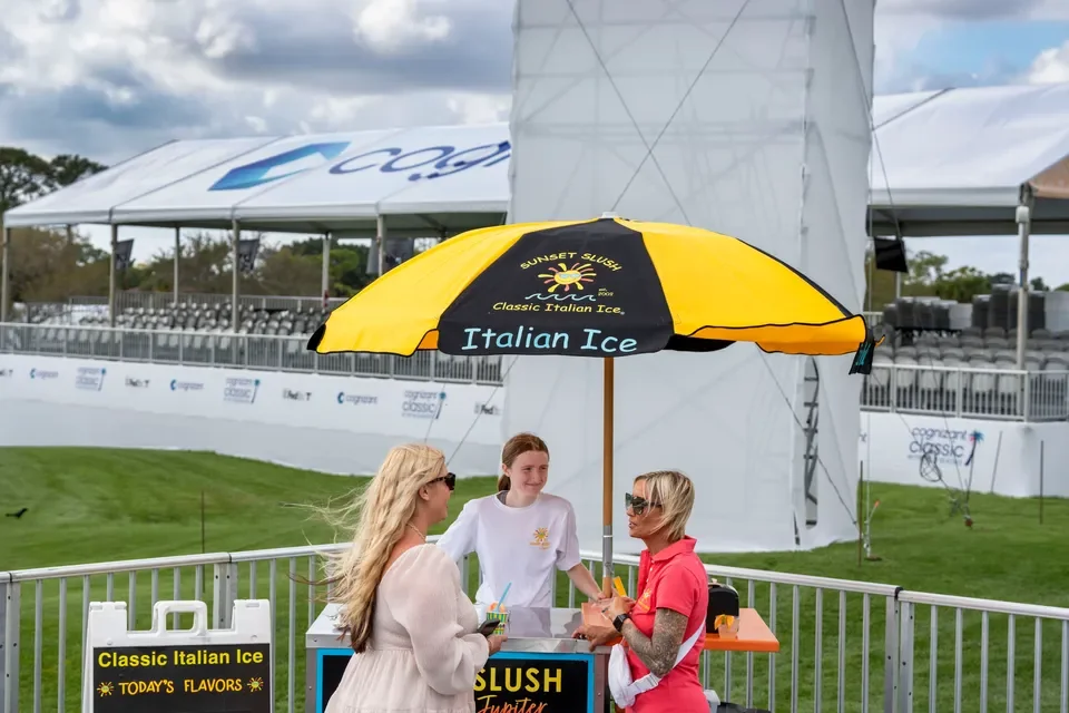 Three women standing near a table with a large yellow and black umbrella labeled "Italian Ice." Behind them is a grassy area, bleachers, and a large white structure at an outdoor event.