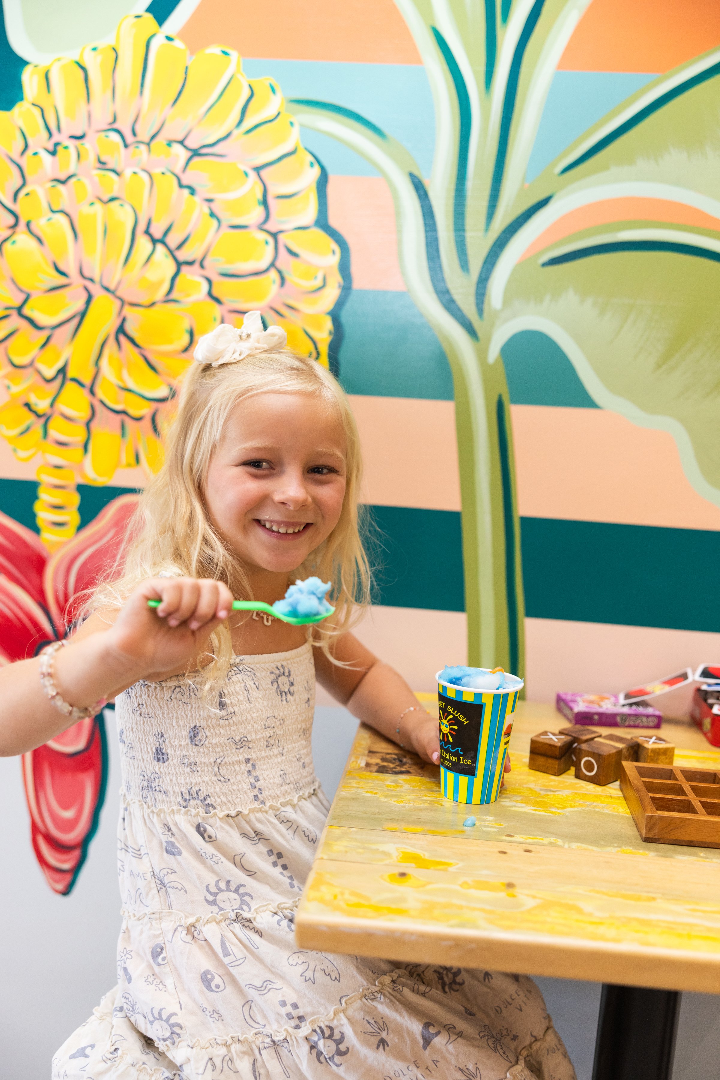 A young girl with long blonde hair, smiling, holding a spoon with blue slush, sitting at a wooden table with a colorful mural of pineapple and leaves in the background.