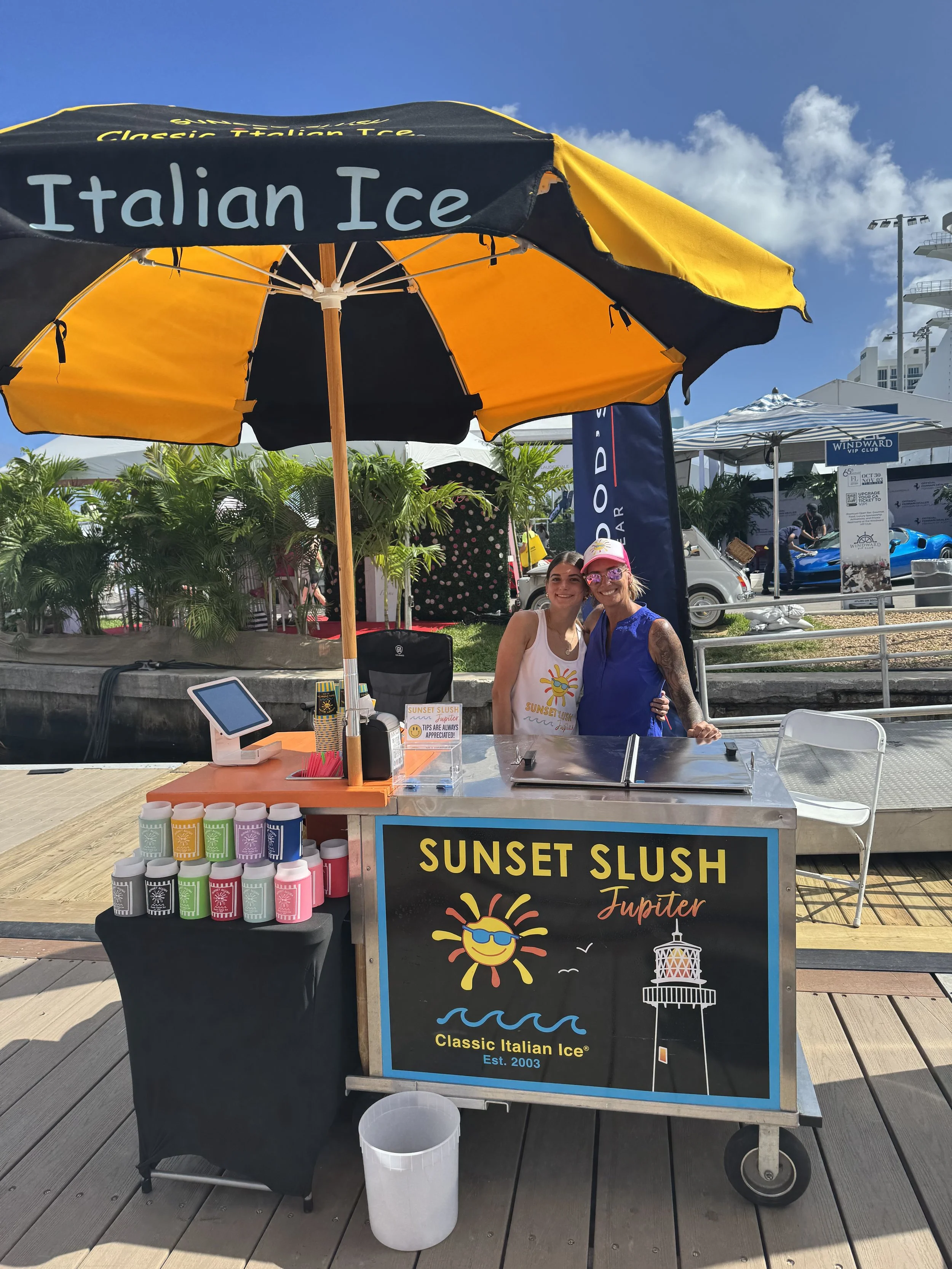 A colorful beachside stand selling Sunset Slush, an Italian ice treat, with two smiling women behind the counter. The stand has a large yellow and black umbrella with "Italian Ice" written on it, and displays small containers of flavored ice, a credit card reader, and a sign promoting the sunset slush product.