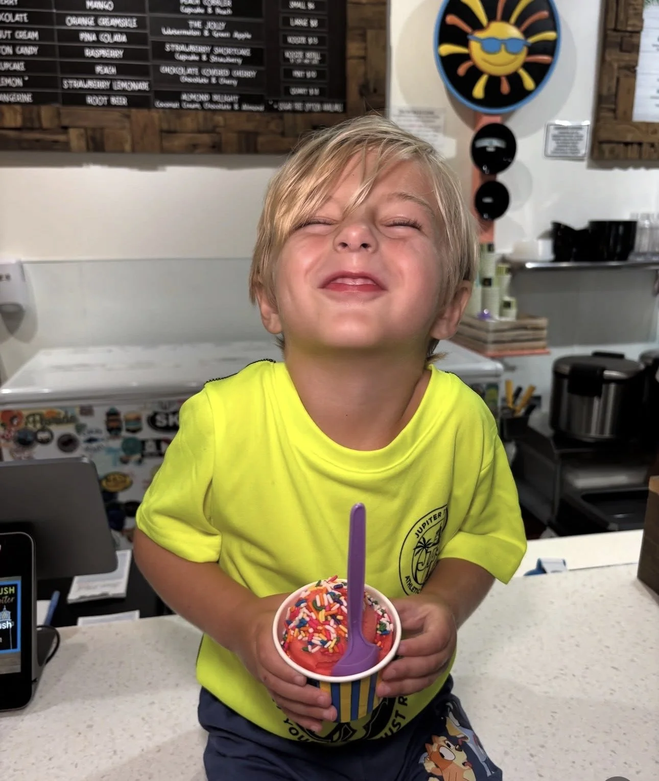 A young boy with blond hair, wearing a bright yellow shirt, is holding a cup of colorful ice cream with sprinkles and a purple spoon. He is smiling joyfully with his eyes closed, standing at a counter in what appears to be an ice cream shop.