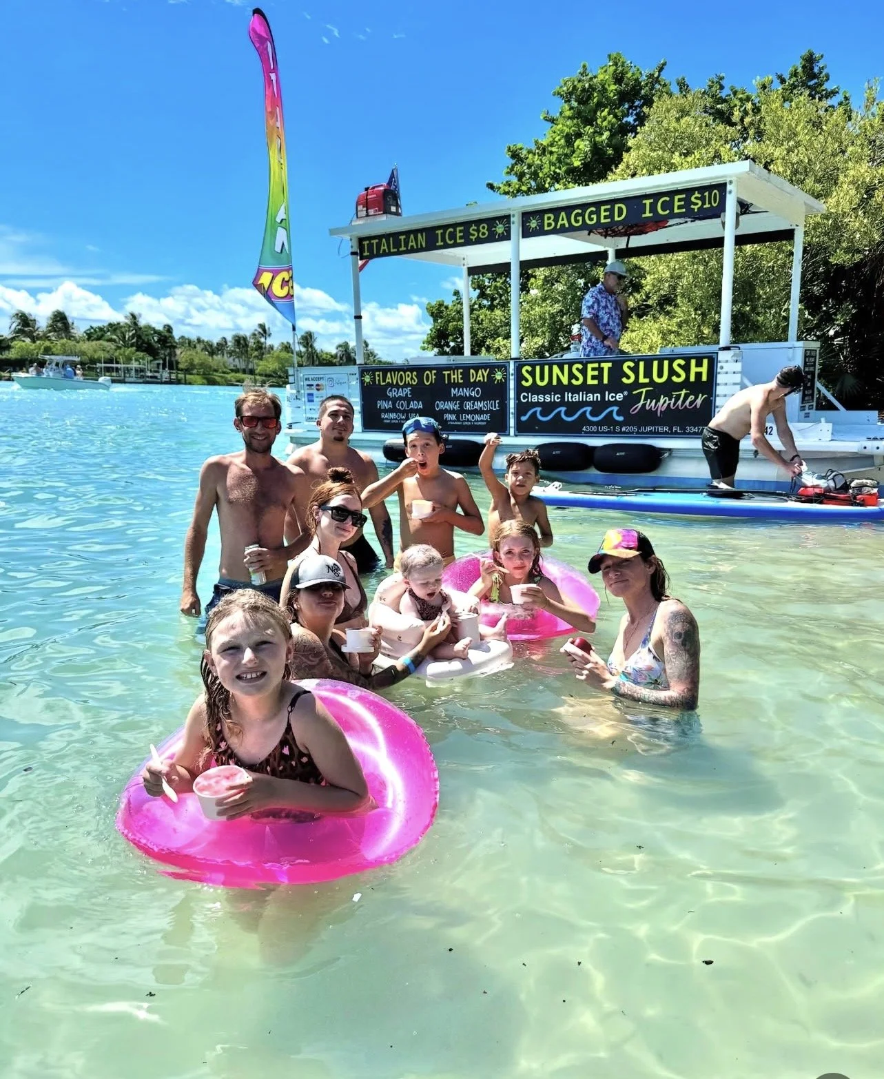 Group of people enjoying ice cream in shallow water near a floating ice cream stand labeled 'Sunset Slush' with flavors and prices, under a bright sunny sky.