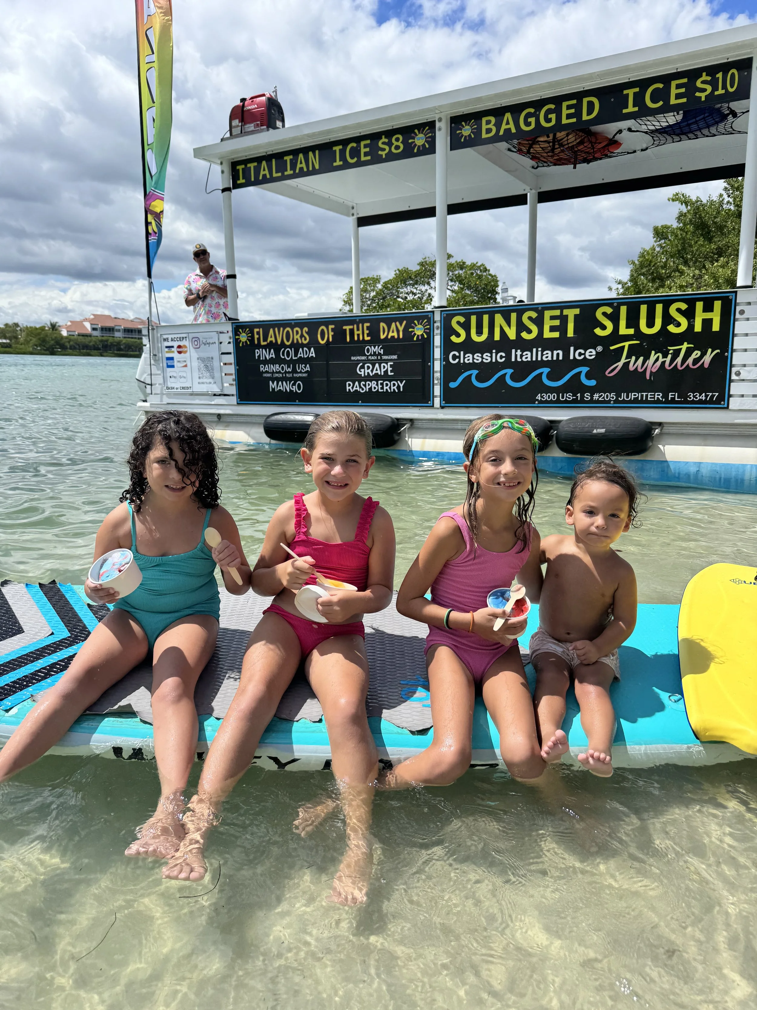 Four children sitting on a paddleboard in shallow water, holding ice cream, at a boat with signs advertising ice cream flavors and drinks, with a cloudy sky in the background.
