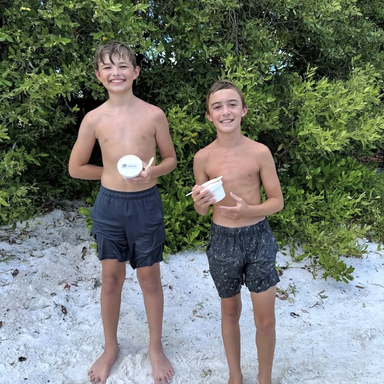 Two smiling boys in swimsuits holding ice cream cups standing on sandy ground in front of green bushes.