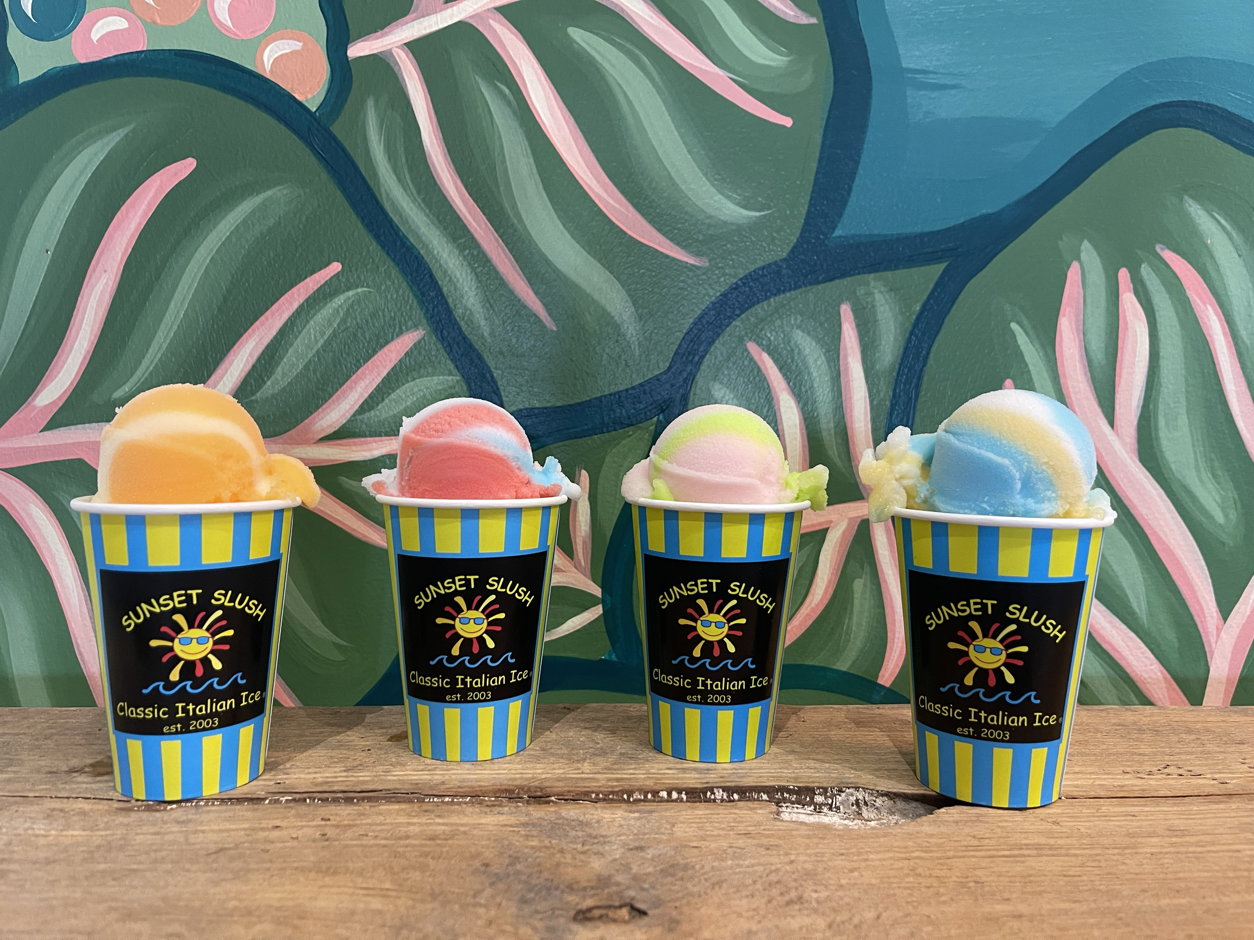 Four cups of colorful Italian ice with different flavors, placed on a wooden surface against a green and pink abstract mural background.