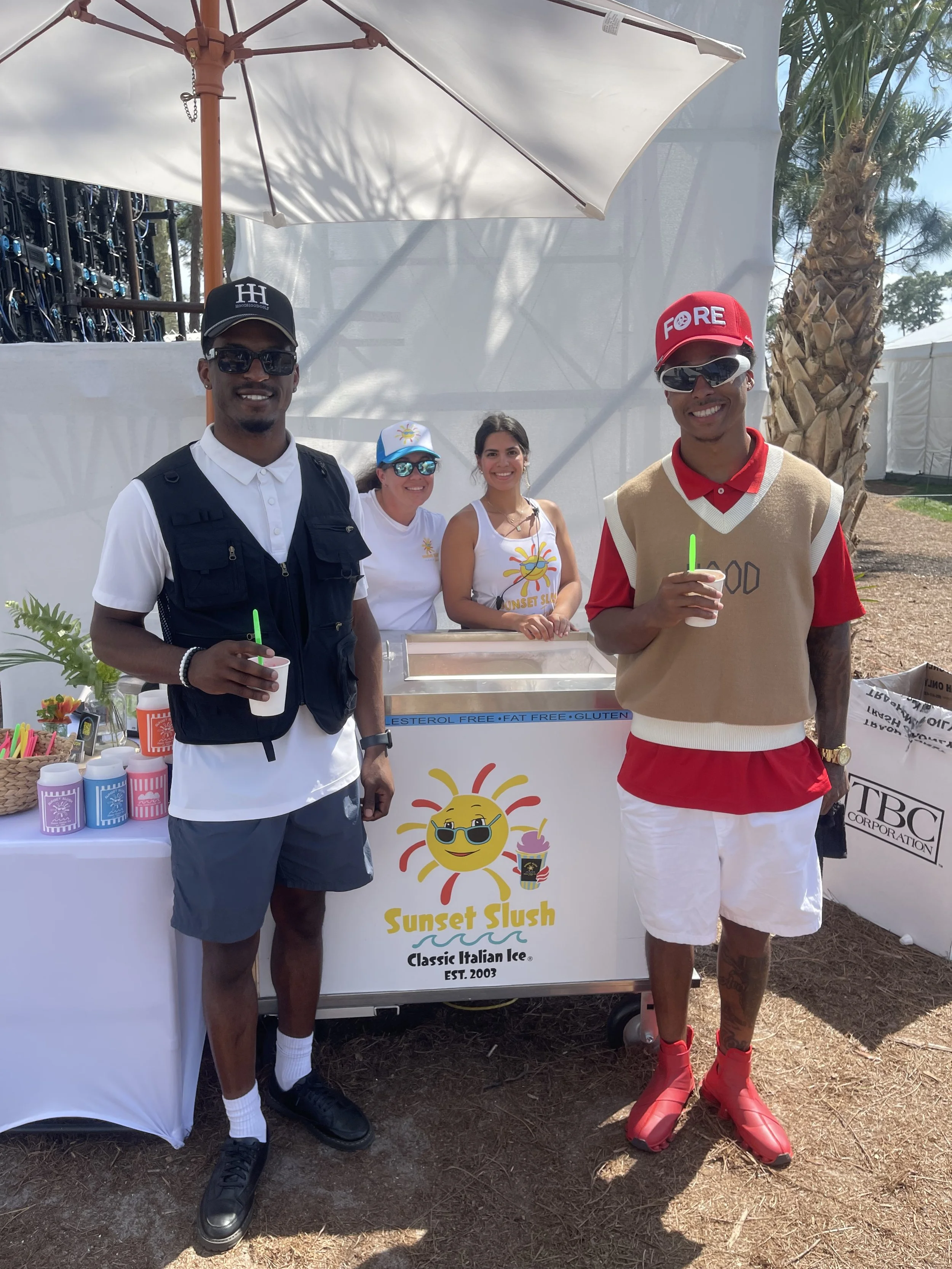 Four people standing behind a slush stand, smiling and holding drinks. Two men in the front, one wearing a black cap, white shirt, and gray shorts, and the other wearing a red cap, beige vest, and white shorts. Two women are behind the stand, one wearing a white tank top and the other in a white T-shirt. The stand has a sign reading 'Sunset Slush,' with a smiling sun logo wearing sunglasses, and various colorful cups and straws on the table.