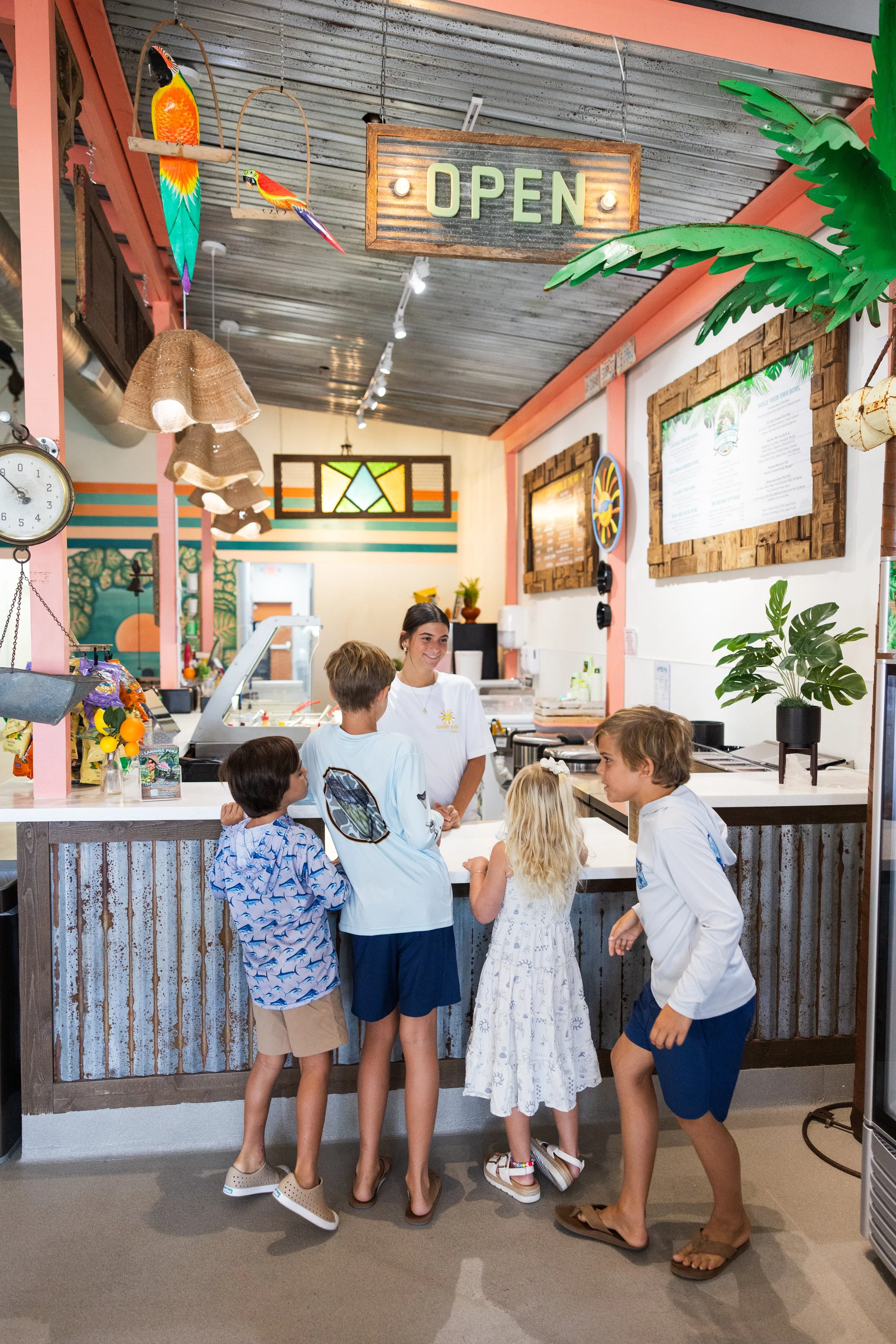 A group of children at a counter in a colorful, tropical-themed cafe or restaurant, with a smiling female staff member taking their orders, and an 'Open' sign hanging from the ceiling.