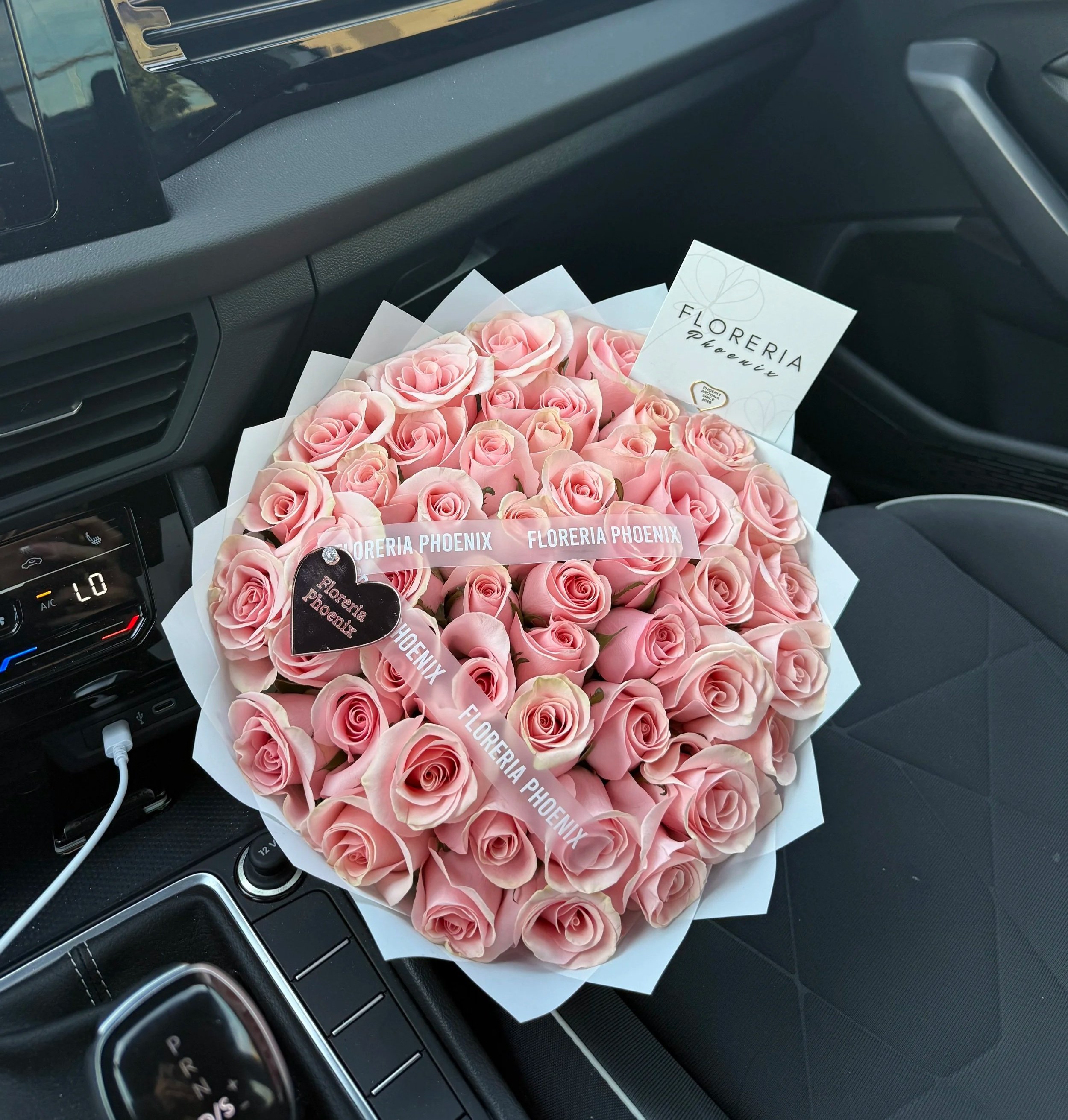 A large bouquet of pink roses with a business card from Floreria Phoenix placed on top of the bouquet inside a car. The car's dashboard and center console are visible.