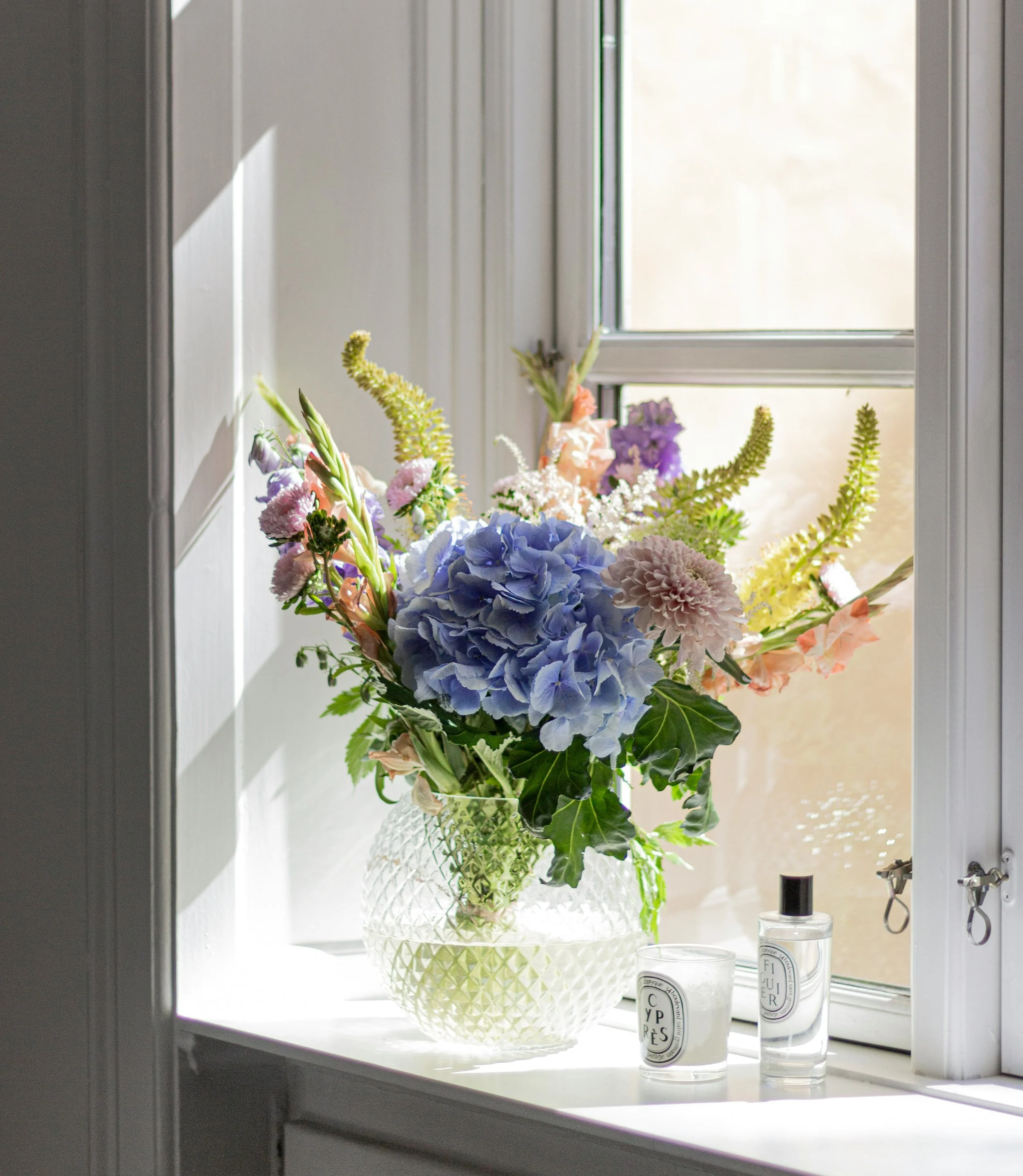 A window with a white windowsill holding a decorative white basket filled with pastel-colored flowers and green leaves. Two candles in glass containers labeled with chic black and white labels sit beside the basket. The scene is lit by natural light, creating a bright indoor setting.