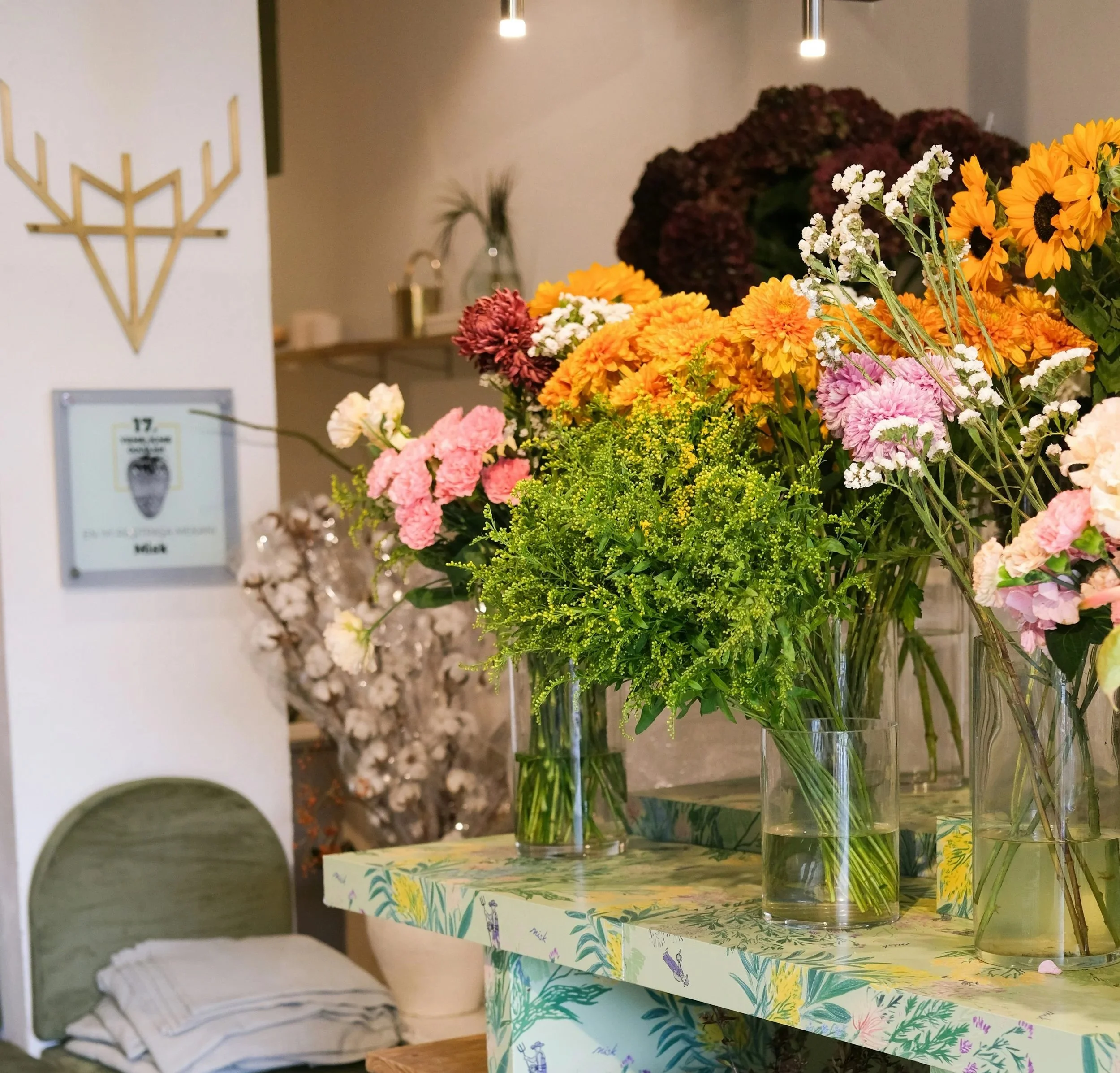 A colorful assortment of fresh flowers in tall glass vases on a table, with a decorative white and gold menorah on the wall in the background.