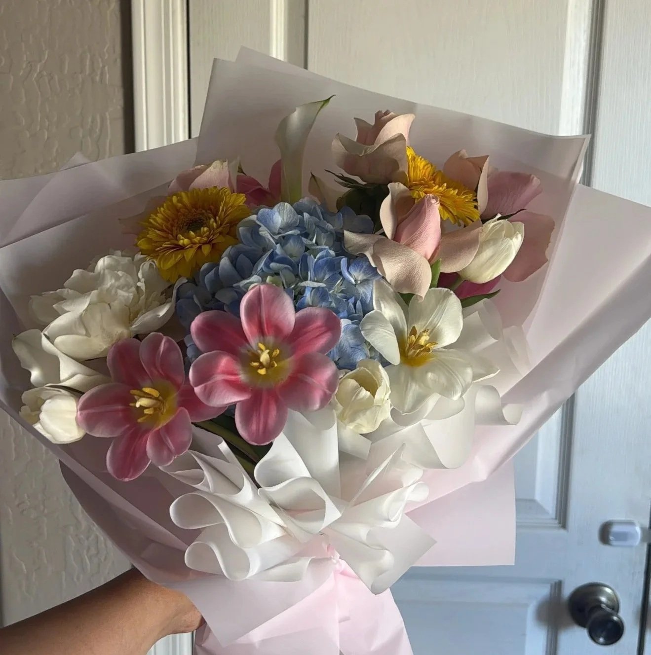 A hand holding a bouquet of mixed flowers wrapped in pink and white paper, including pink tulips, yellow daisies, white calla lilies, and blue hydrangeas.